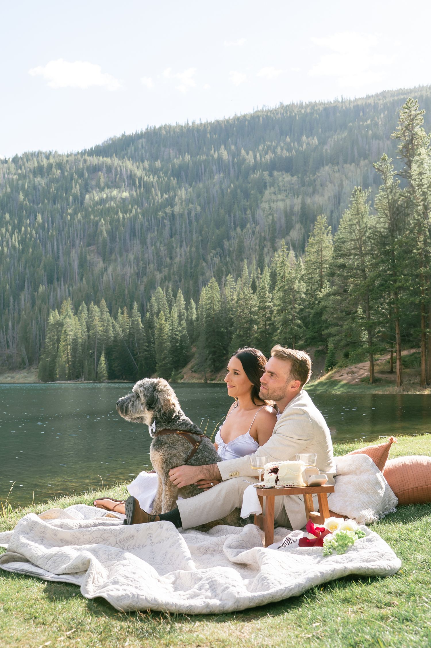 Couple sitting on blanket by mountain lake with dog during outdoor picnic engagement session in scenic forest setting.