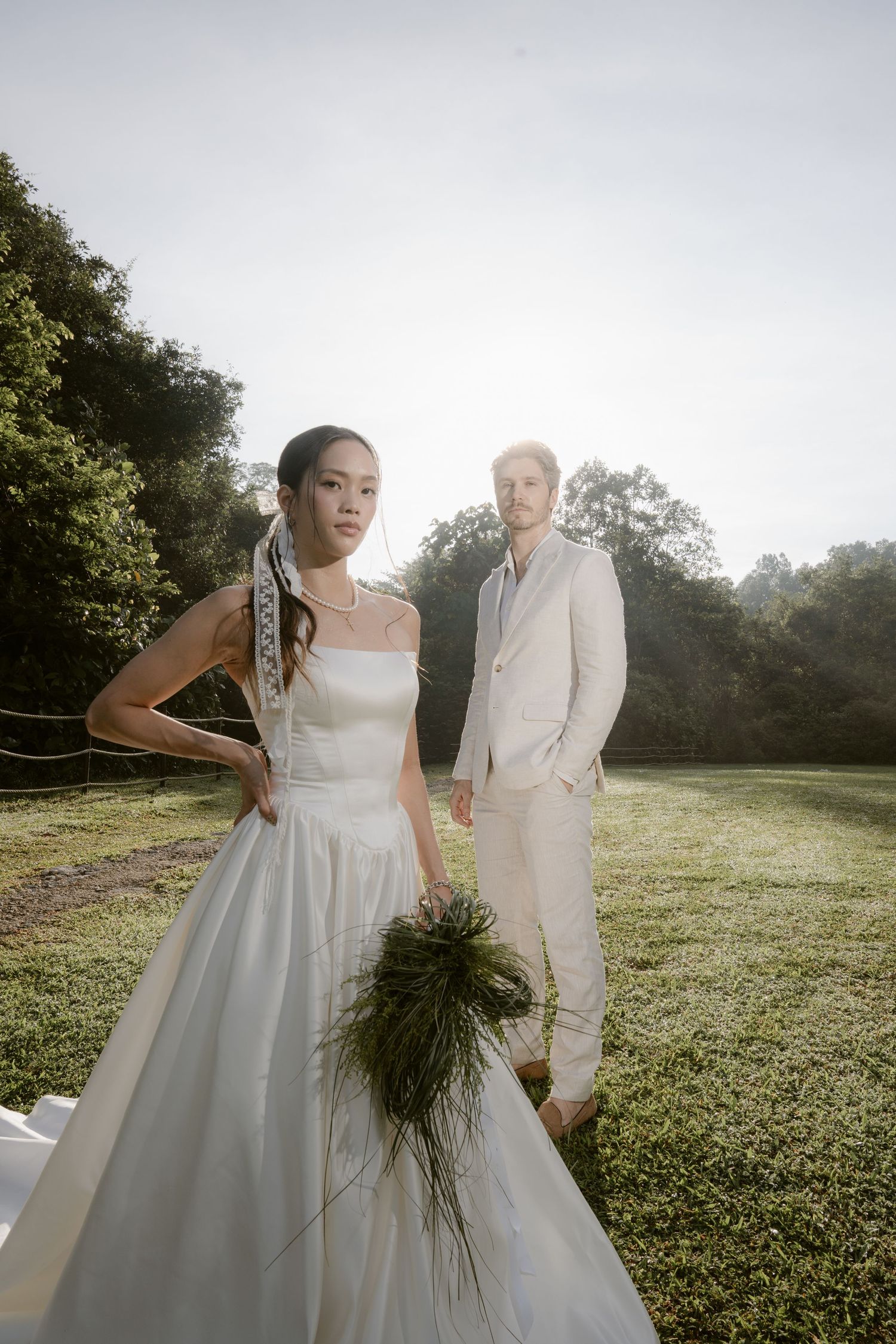 Bride in strapless white gown holds greenery bouquet as groom in beige suit stands behind her outdoors.
