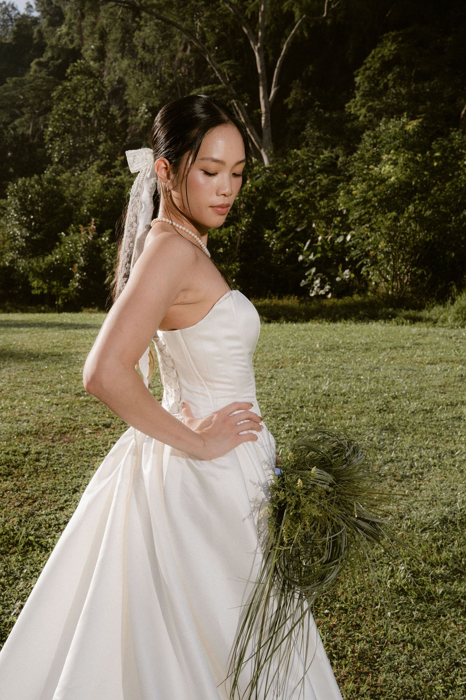 Bride in strapless white satin gown with veil holds greenery bouquet in sunlit garden setting.