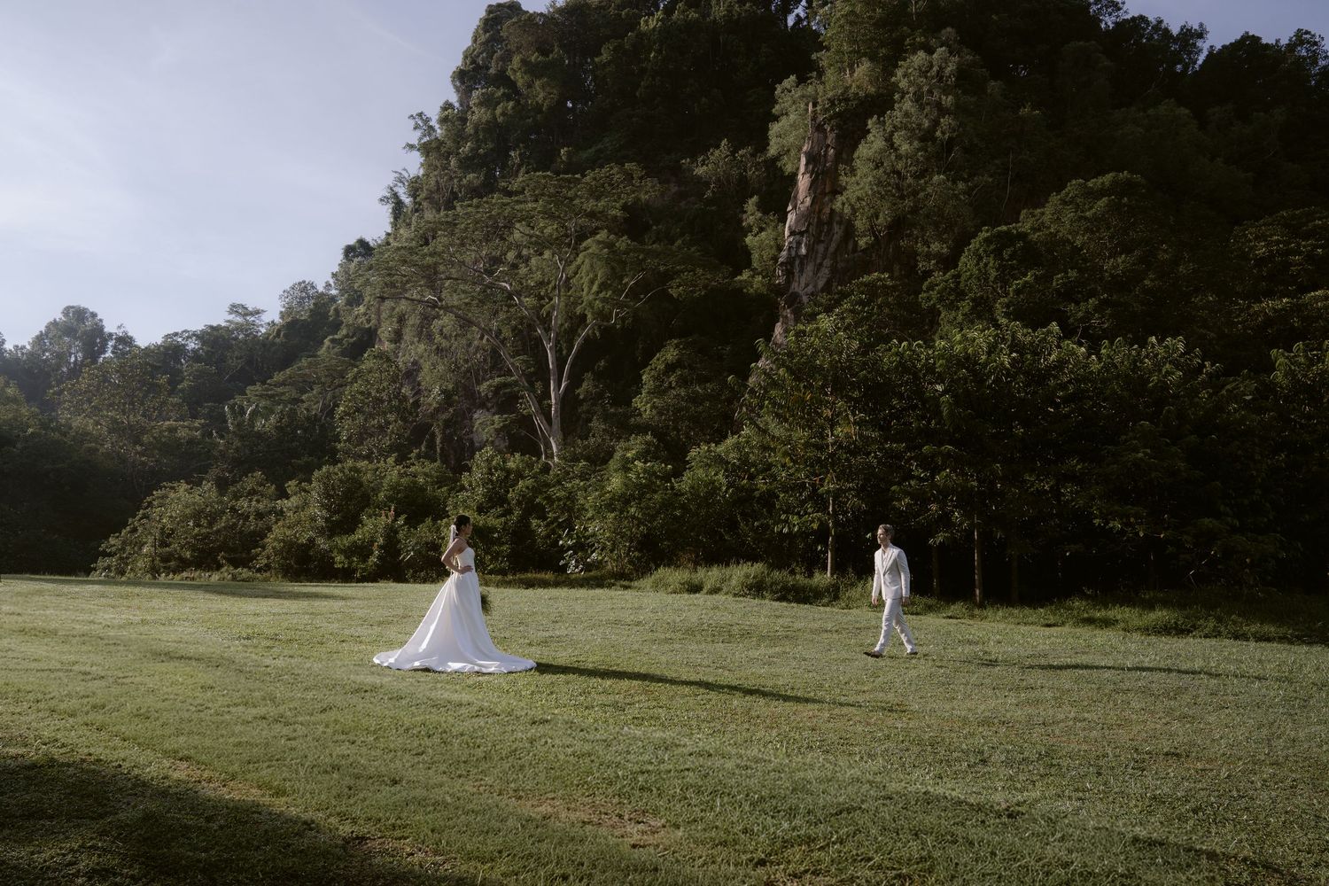 Bride and groom stand apart on lush green lawn with dramatic forested rocky cliff in background.