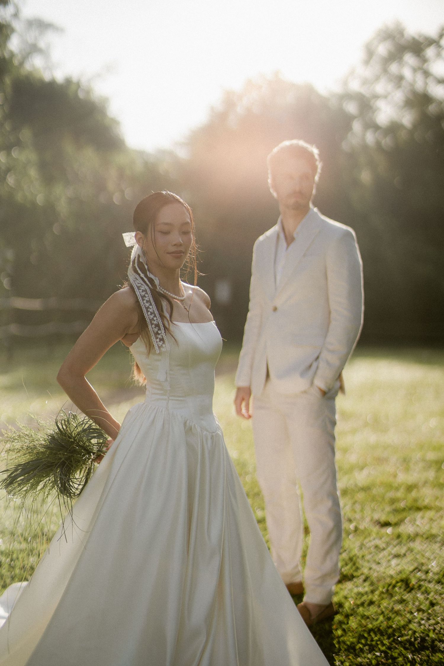 Bride in white gown with lace sash and groom in cream suit in golden sunlit field.