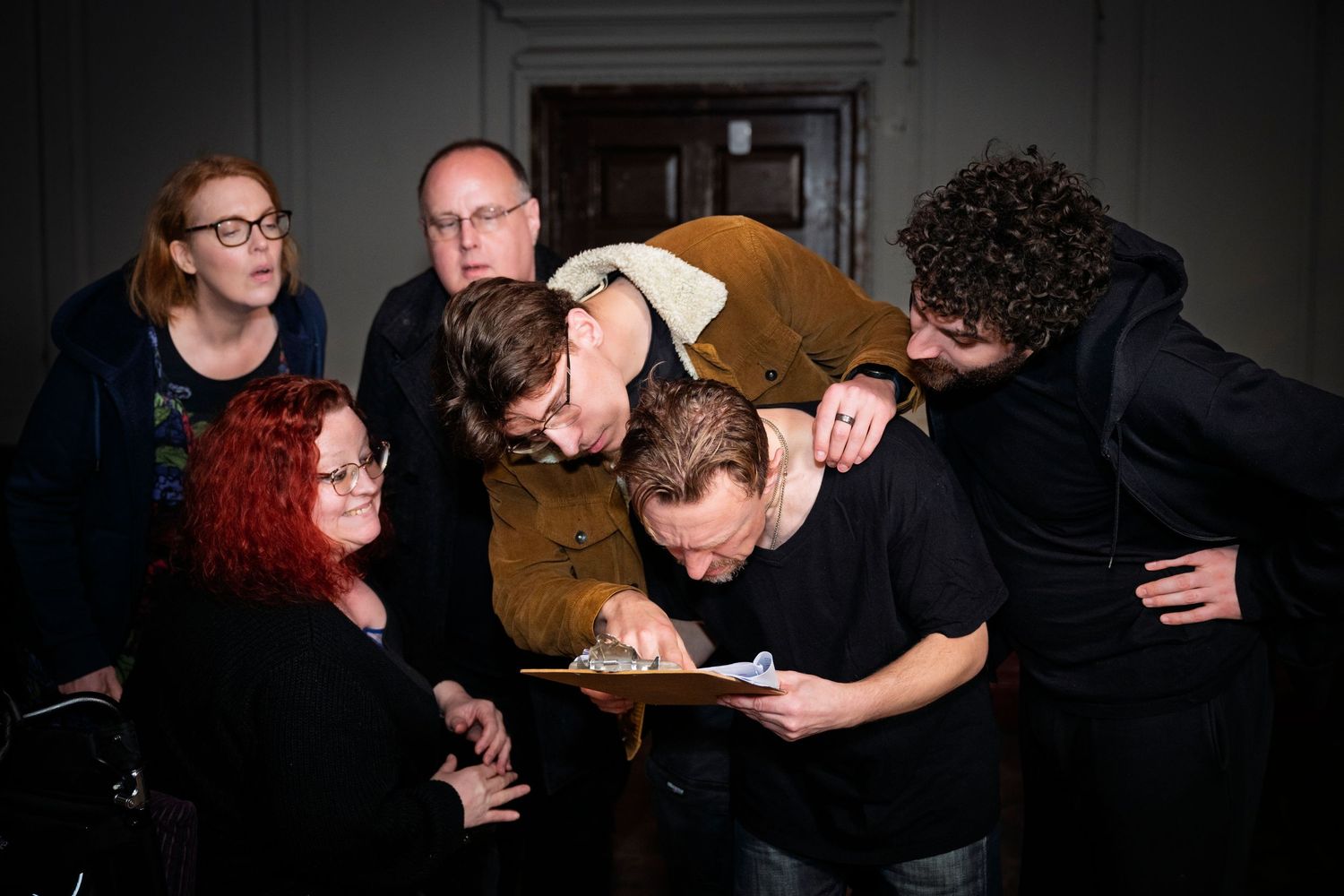 Group of six adults huddled together examining a document in a dimly lit room.