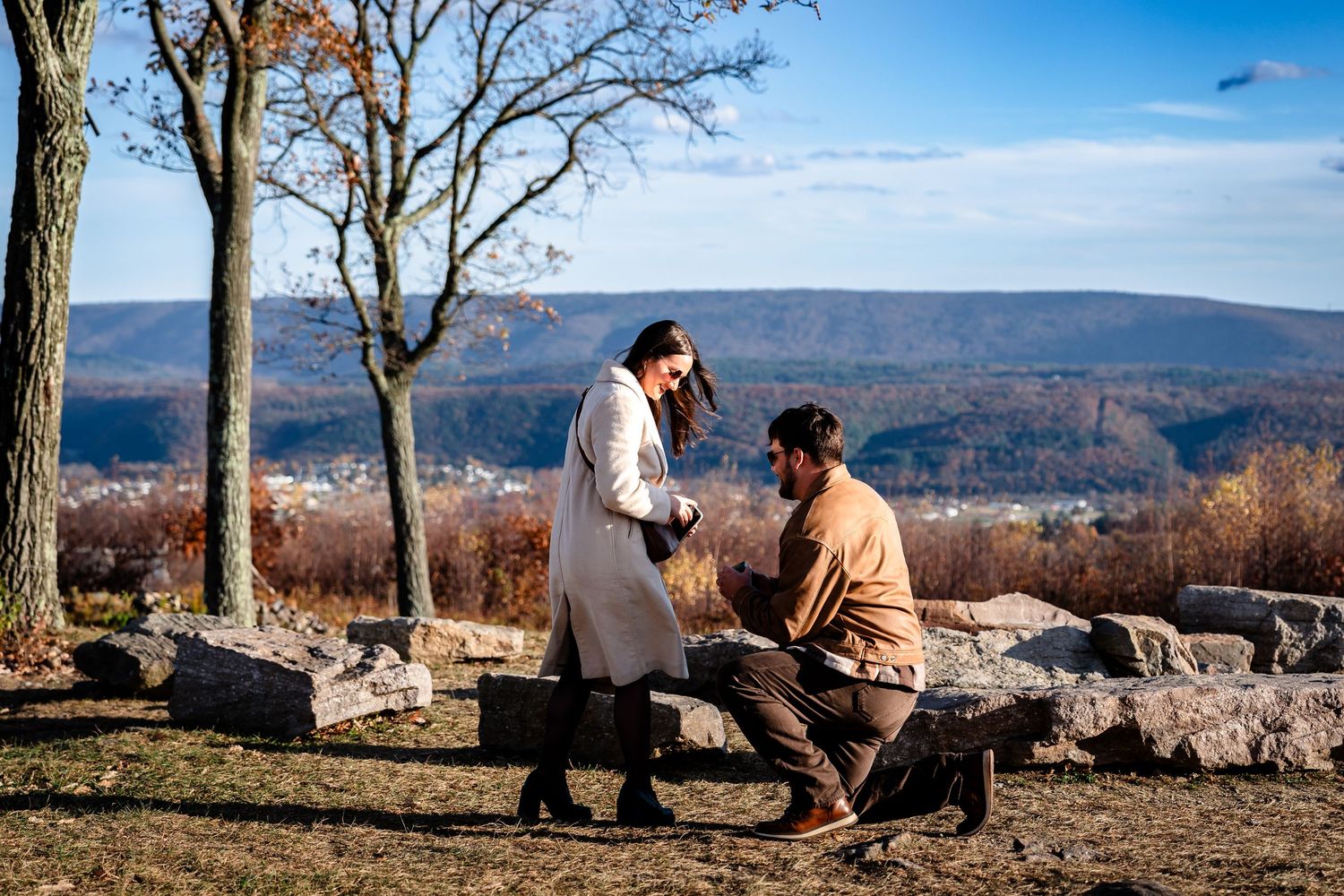Man kneeling to propose to woman on rocky hilltop with scenic mountain valley view in autumn.