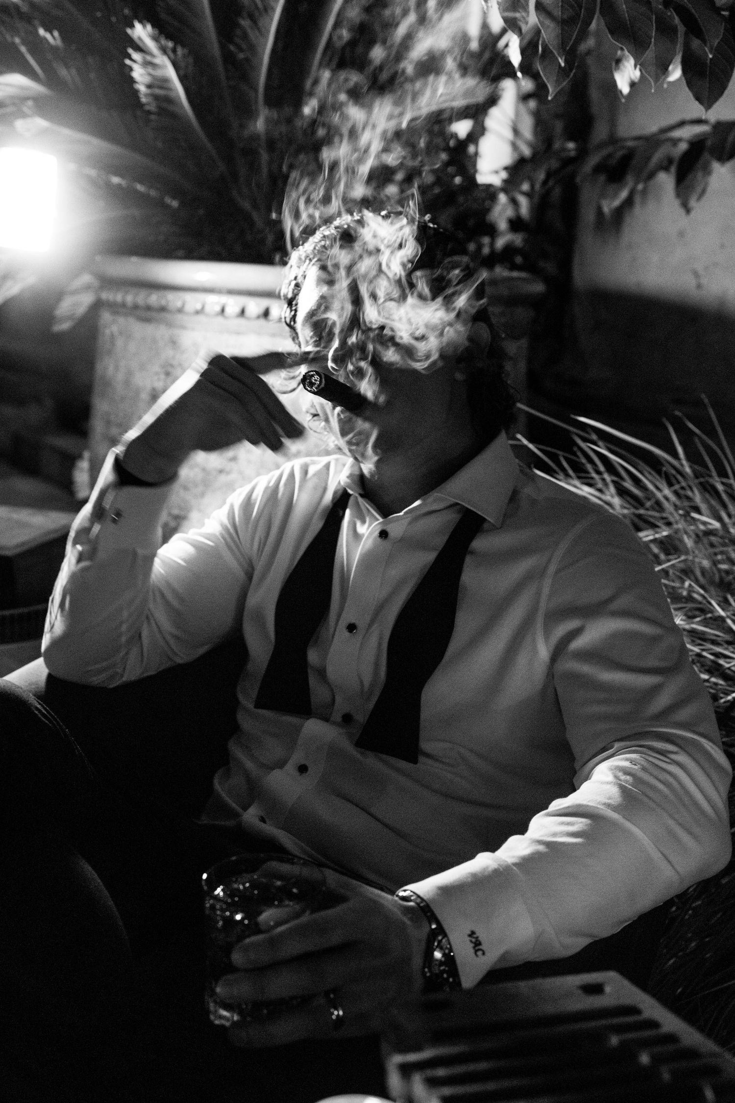 Black and white photo of a man in a white shirt and tie smoking a cigar while holding a drink outdoors.