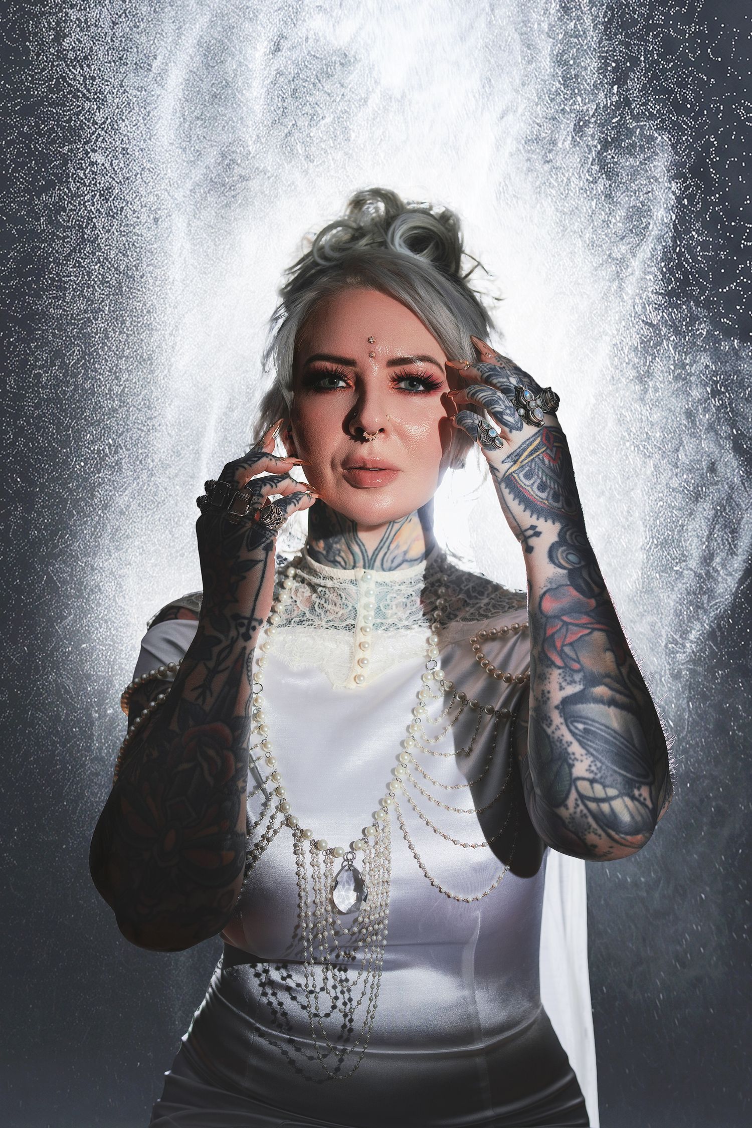 Tattooed woman with gray hair wearing pearl necklace and lace gloves posing dramatically with powder explosion backdrop.