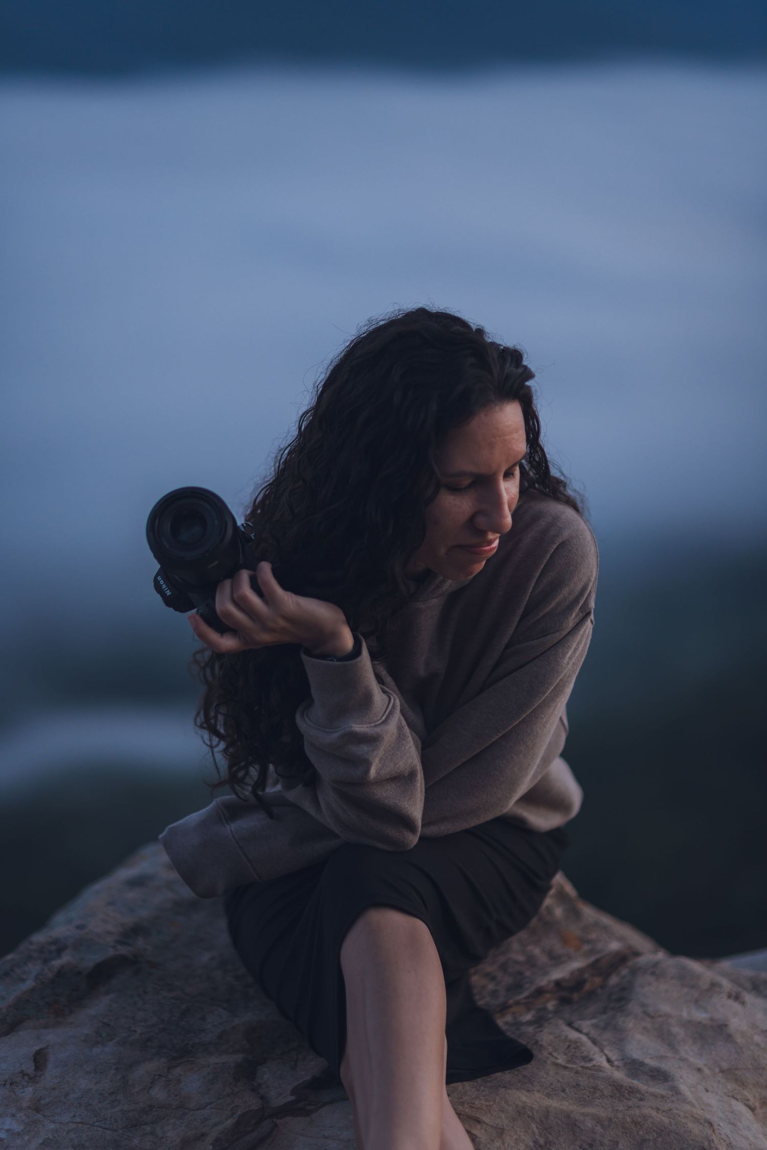 A photographer takes pictures during sunset on rocky coastal cliffs with misty ocean waves in the background.