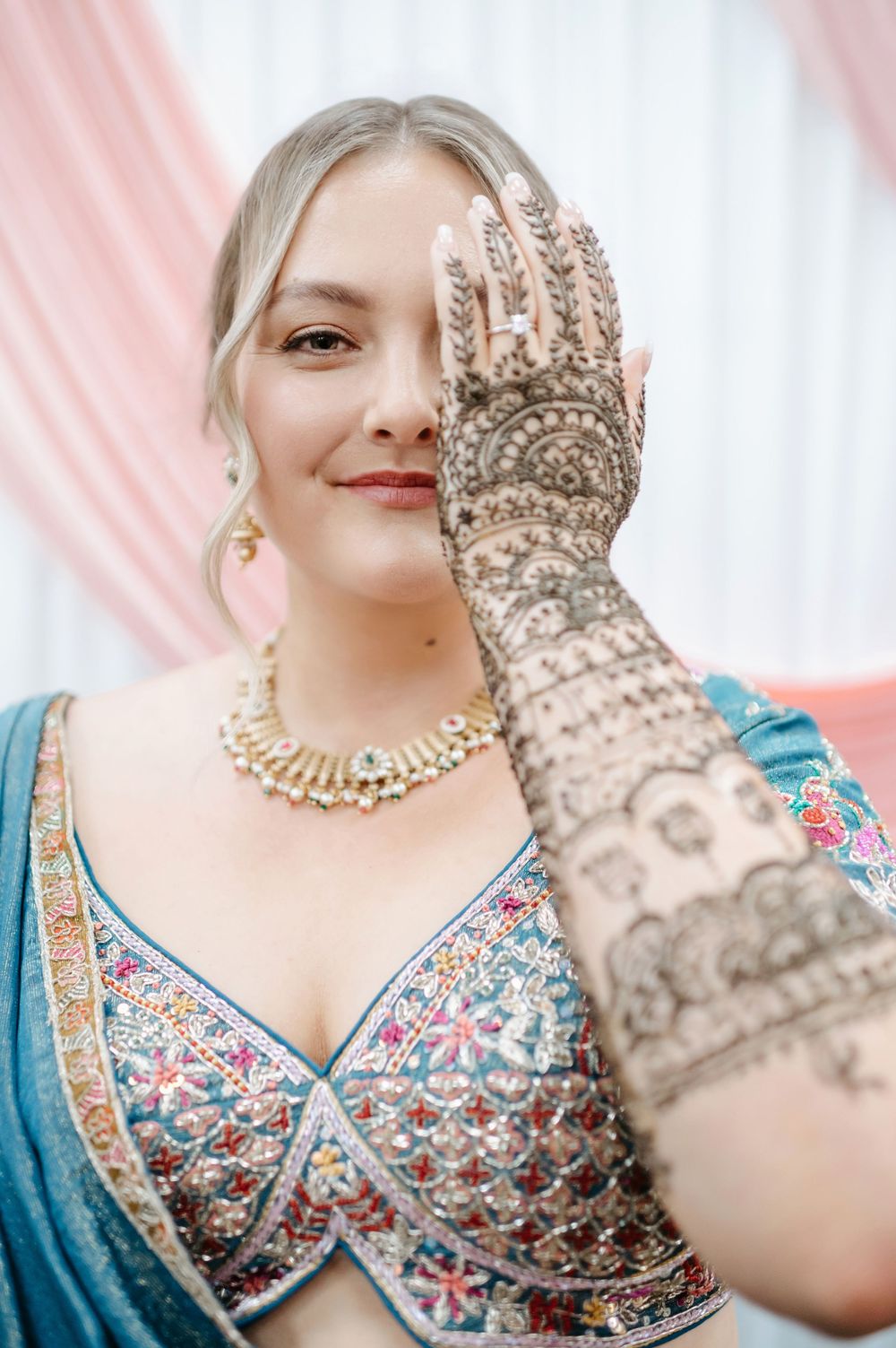 Blonde bride in blue lehenga displays intricate mehndi henna hand art, wearing gold kundan necklace at Indian wedding.