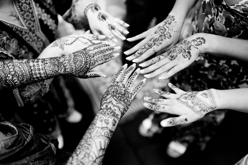Black and white photo of multiple hands adorned with intricate henna mehndi designs at a traditional ceremony.