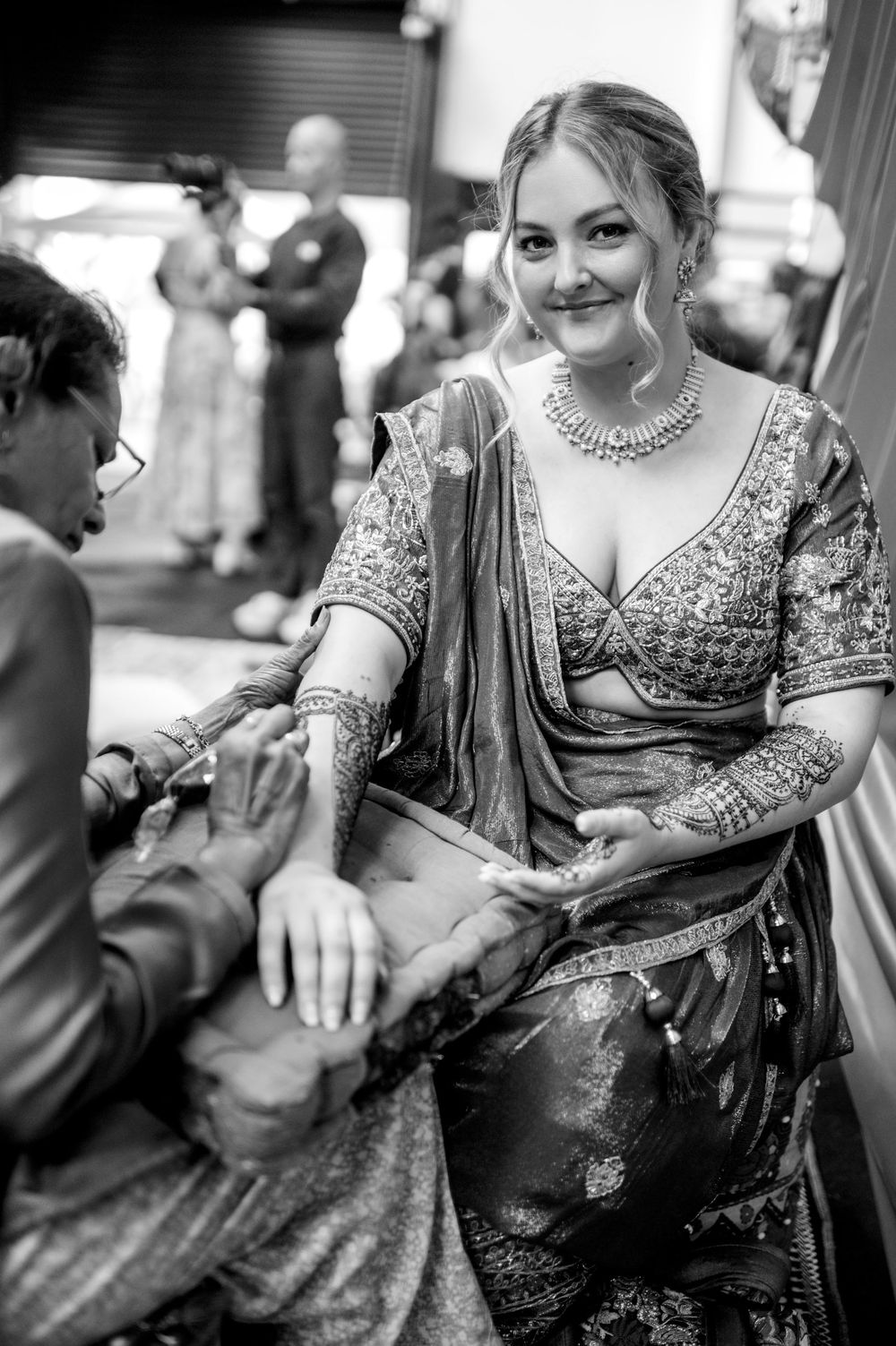 Bride in embroidered lehenga smiles as artist applies mehndi henna to her hand at Indian wedding ceremony.