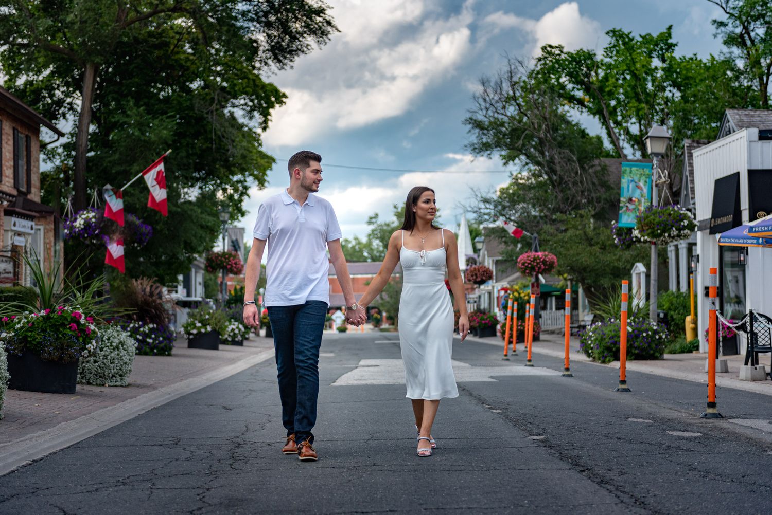 Couple holding hands walking down a charming flower-lined street with Canadian flags on a cloudy summer day.