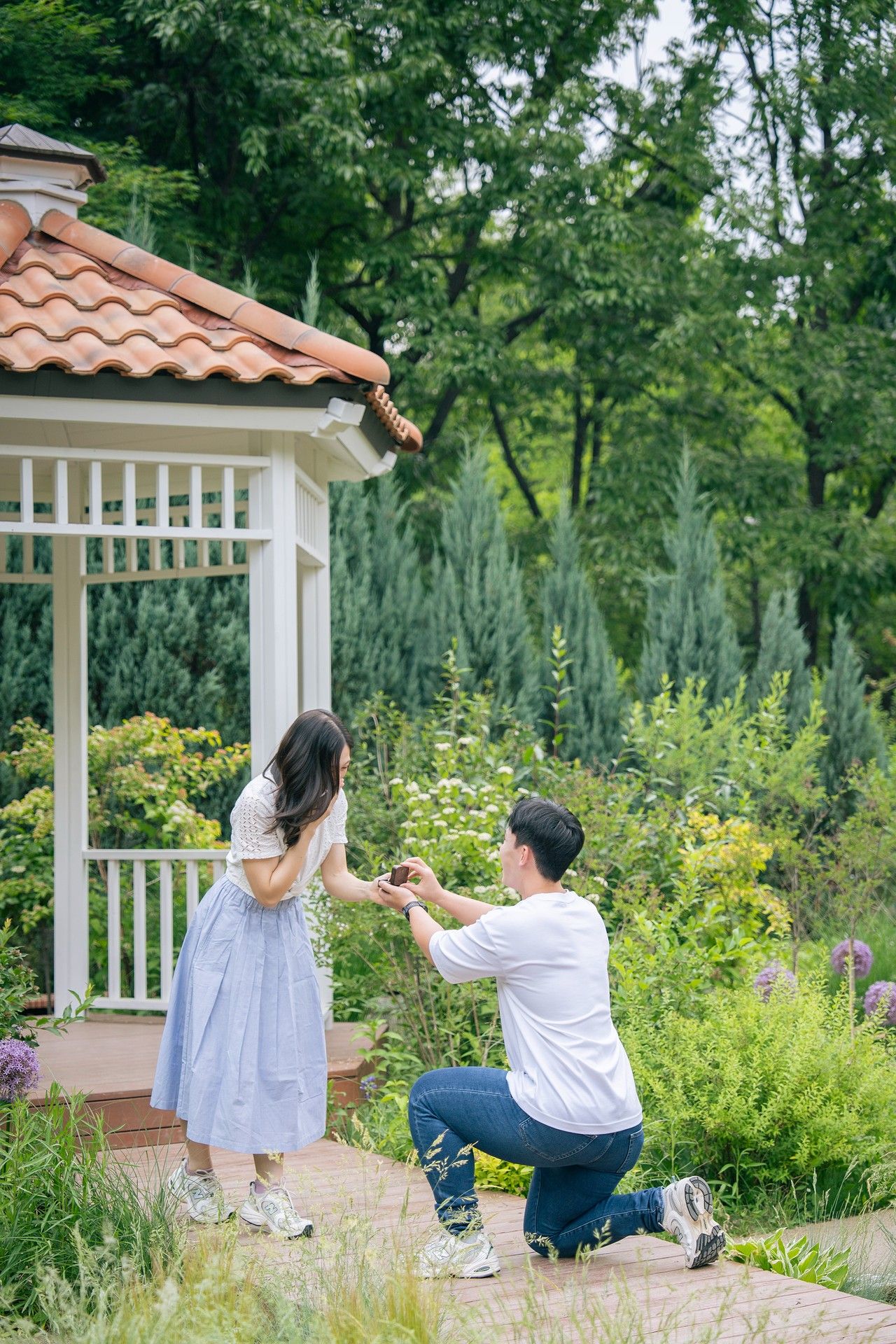 Man proposing on one knee to woman in blue dress near white gazebo in lush green garden.