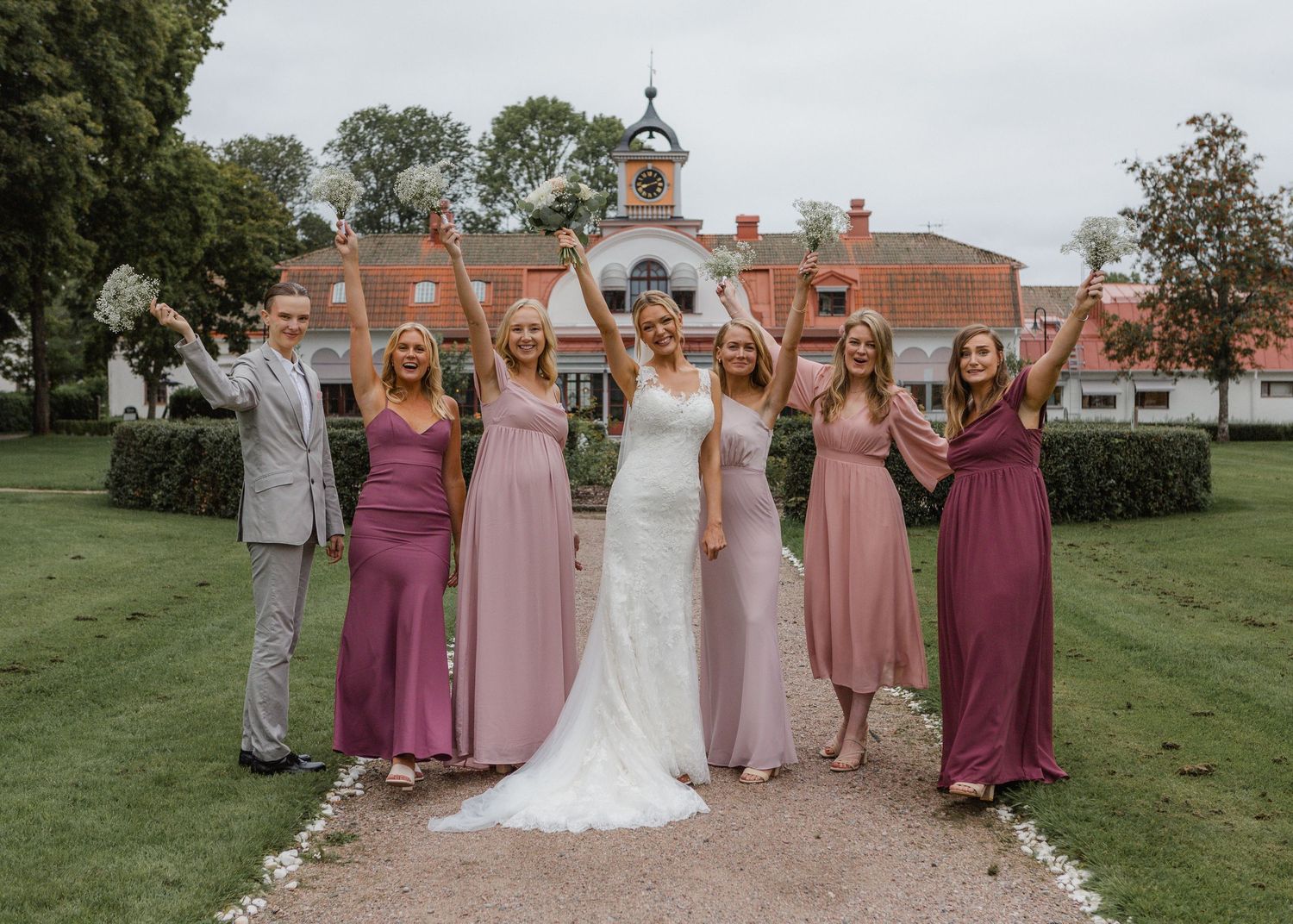Bride in white gown poses with bridesmaids in pink dresses and groomsman outside elegant manor house.