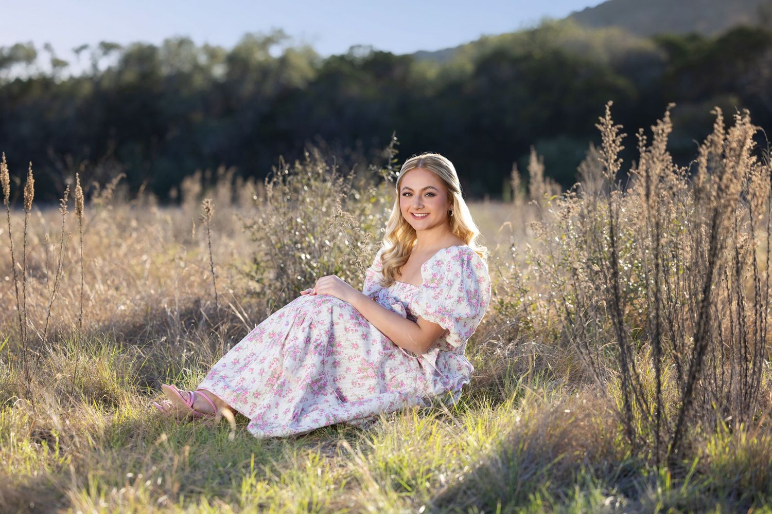 2027 senior graduate in a floral dress sitting in a golden field with trees in the background on a sunny day.