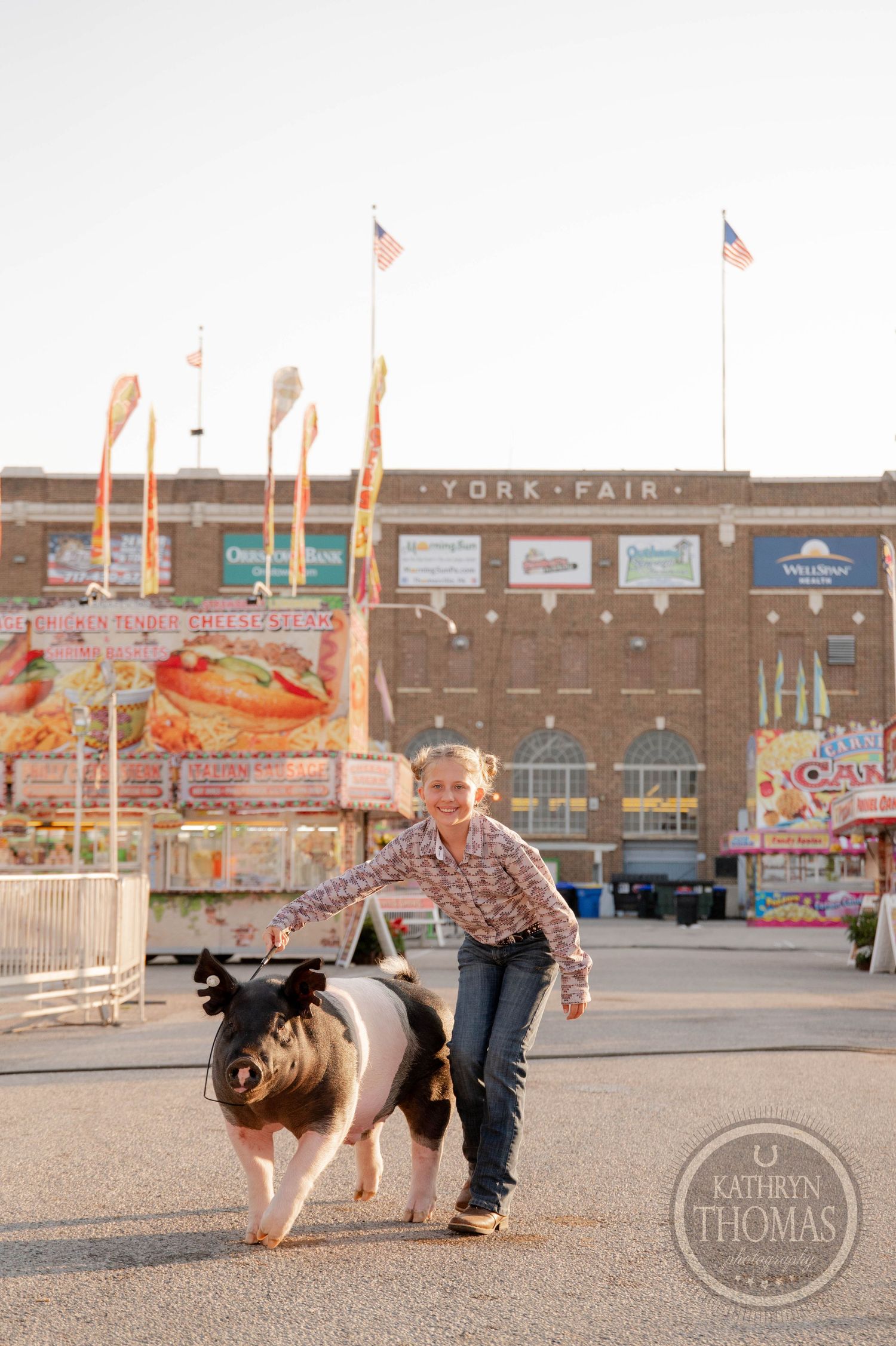 A young girl leads a large pig at the York Fair, with colorful carnival attractions and flags in the background.
