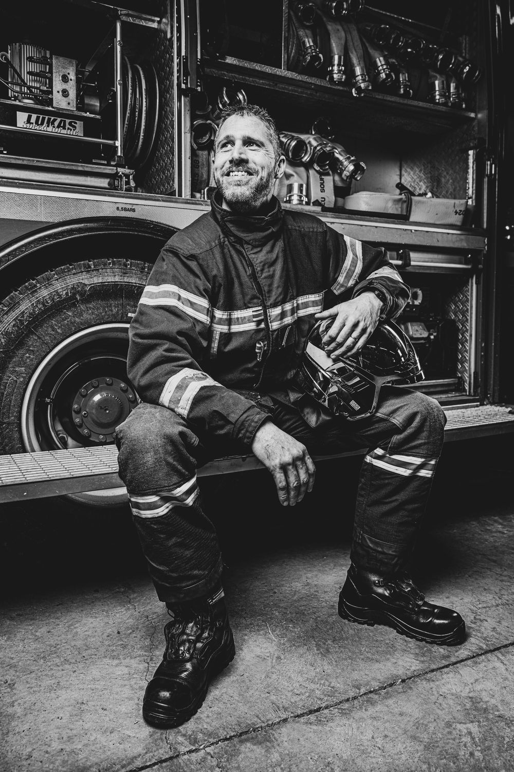 A smiling male firefighter in uniform sits relaxed on a fire truck step in a black and white photo.