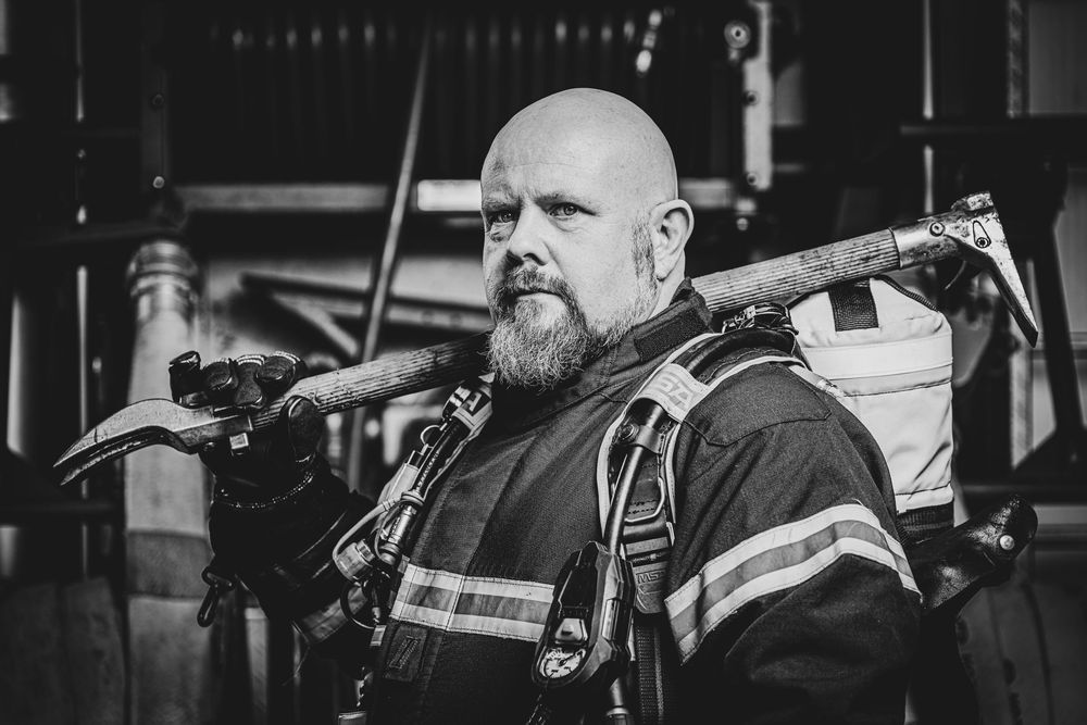 A bald bearded firefighter carries tools over his shoulder in a dramatic black and white photo.