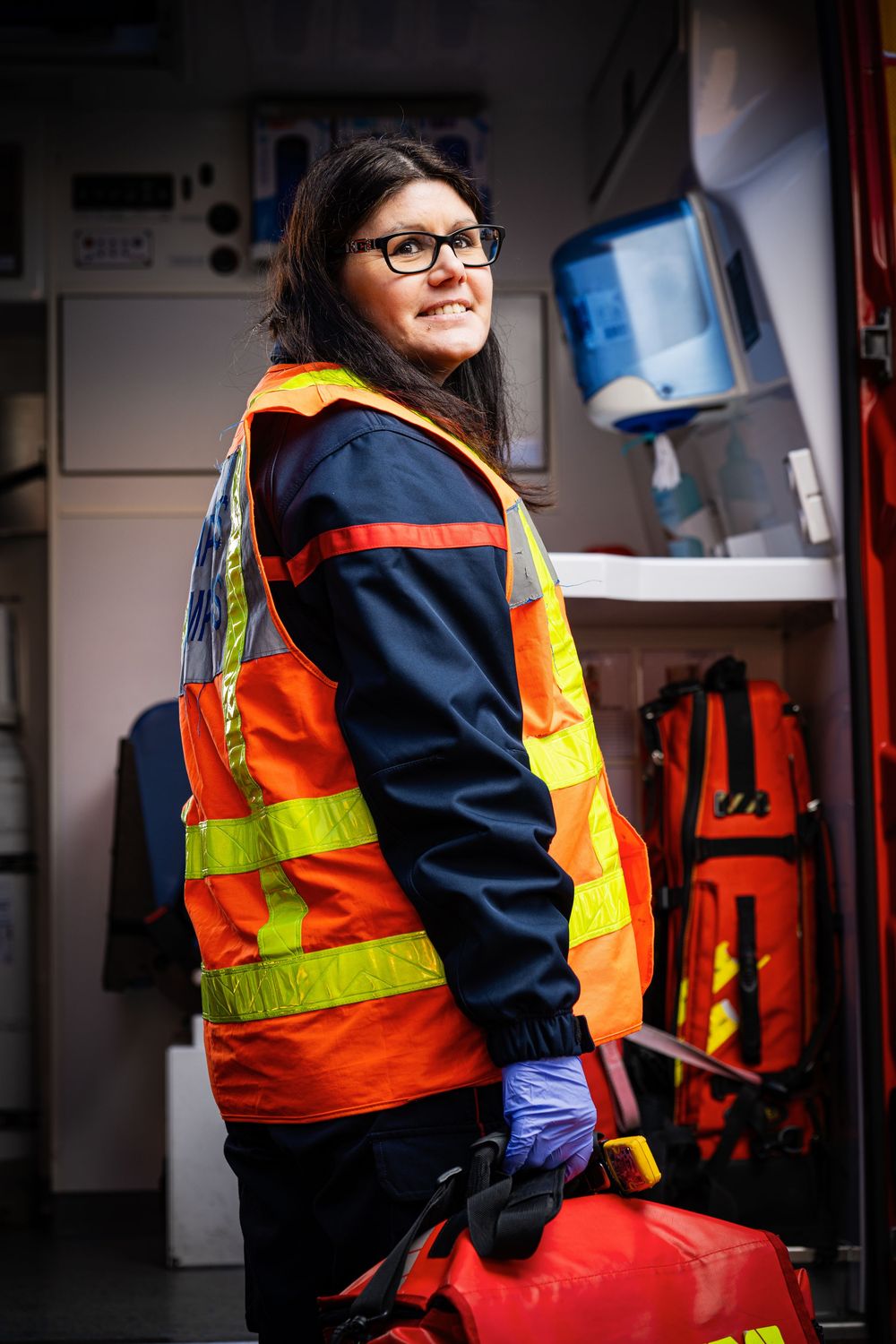 A smiling female paramedic in an orange high-visibility vest stands inside an ambulance holding equipment.