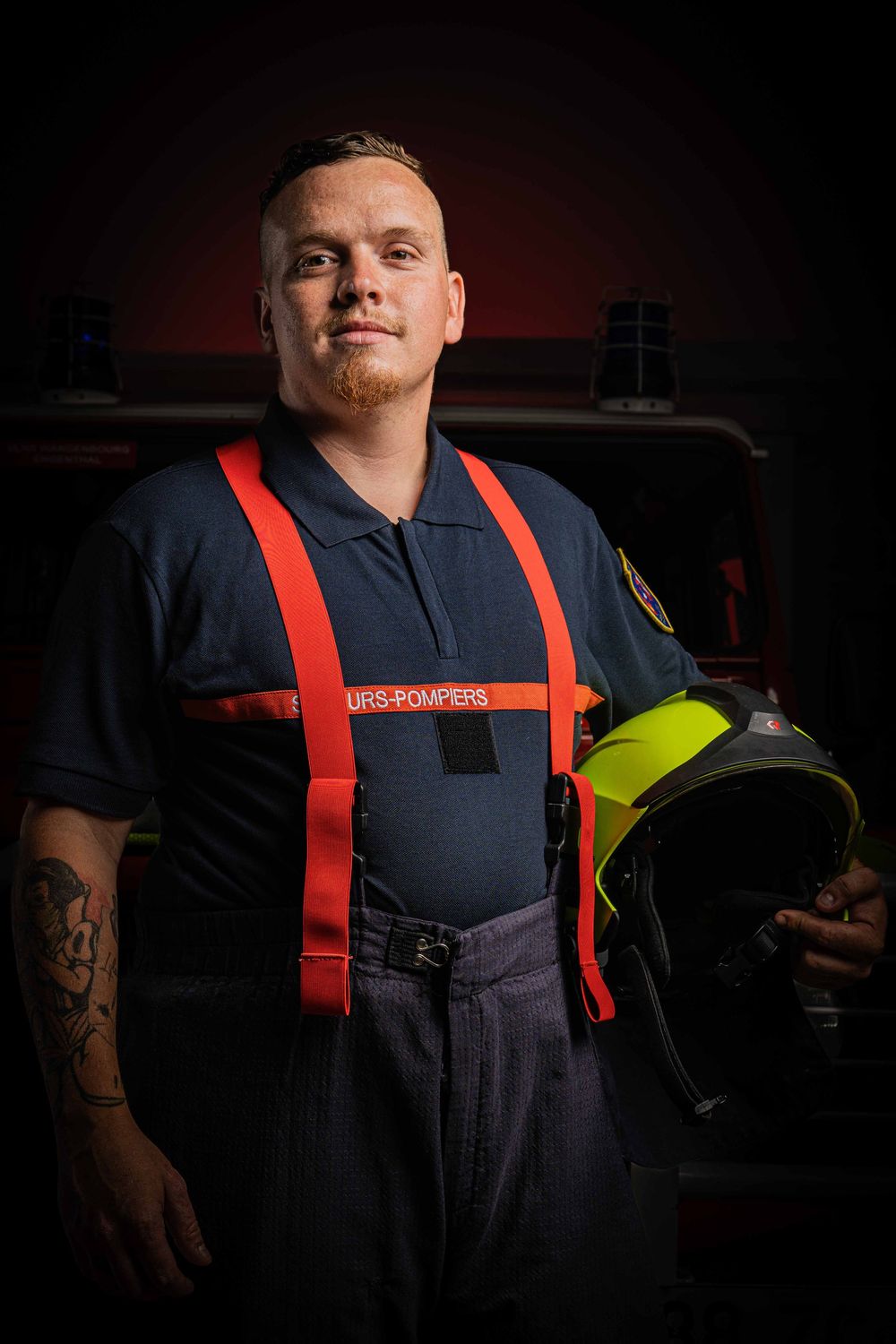 Young male firefighter in uniform with red suspenders holds helmet beside a fire truck.