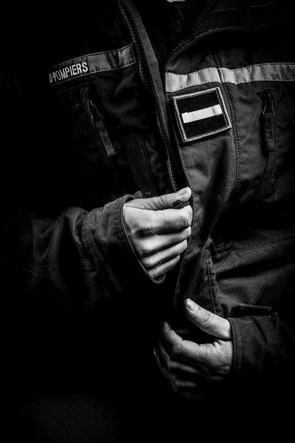 Black and white close-up of firefighter hands gripping a Pompiers jacket with patch.