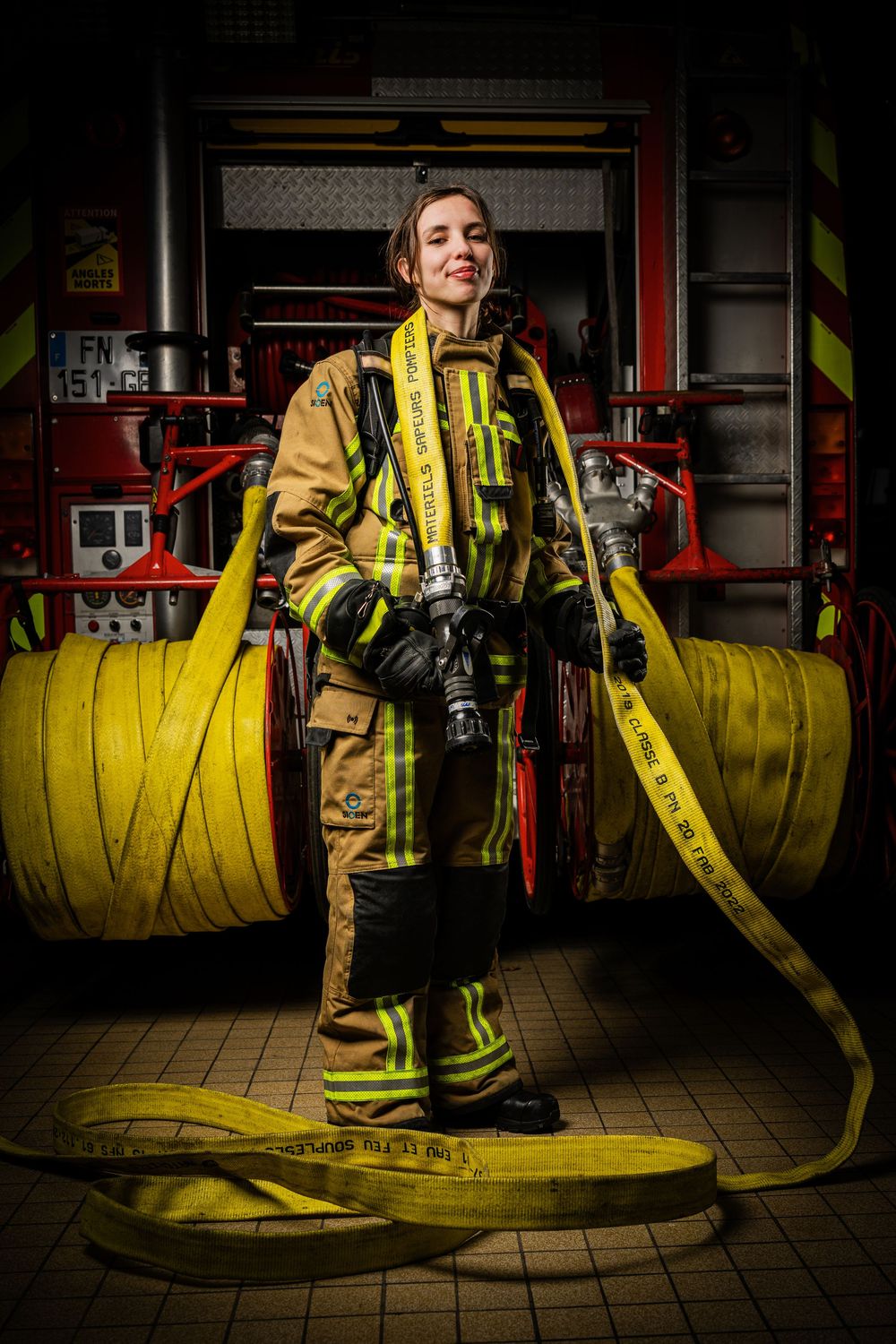 A firefighter arranges yellow fire hoses in a dark fire station with dramatic lighting and equipment visible.