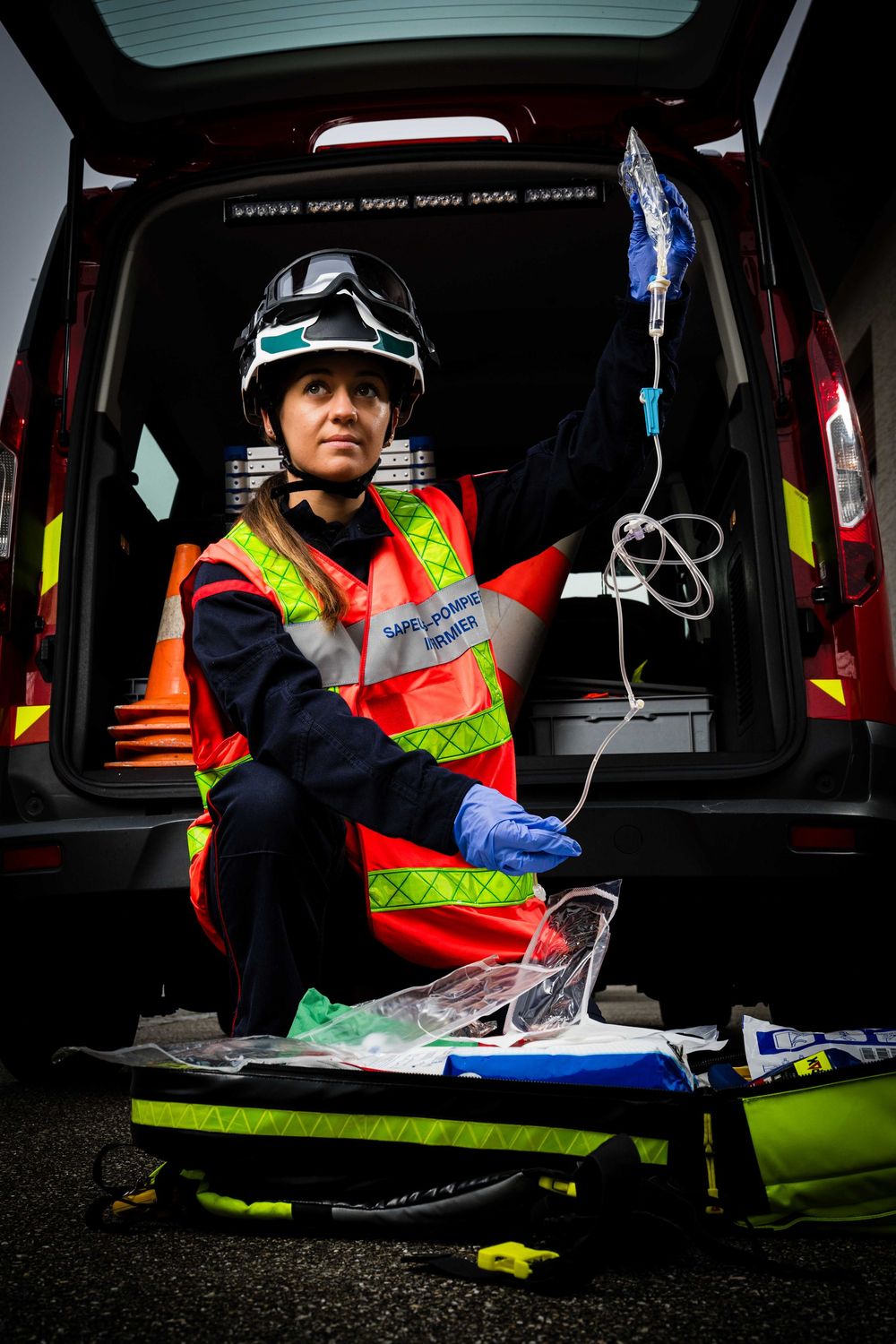 Female paramedic in orange vest holds IV bag while preparing medical equipment at ambulance.