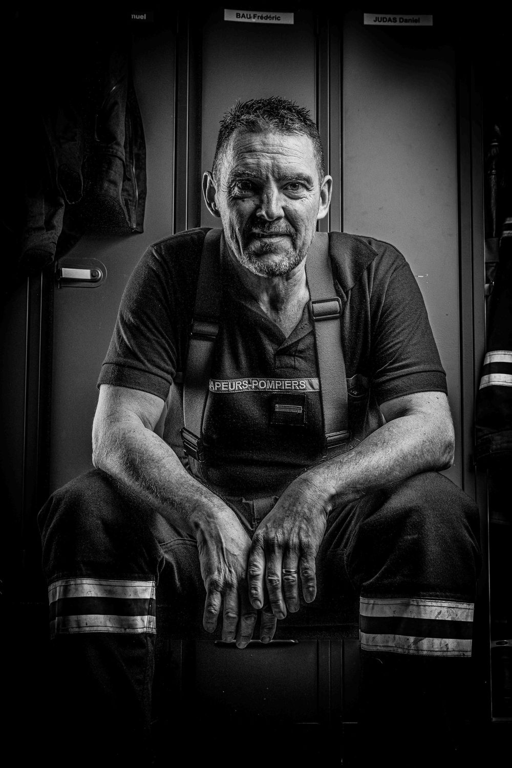 Black and white portrait of veteran firefighter sitting in locker room wearing suspenders.