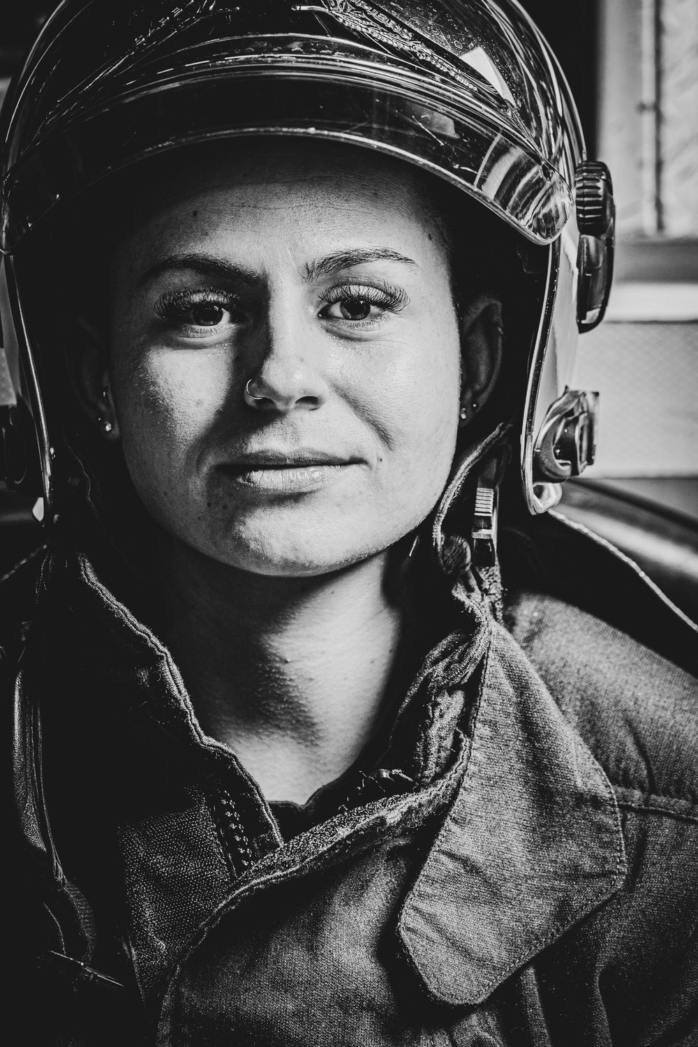 A female firefighter in full helmet and gear poses for a striking black and white close-up portrait.