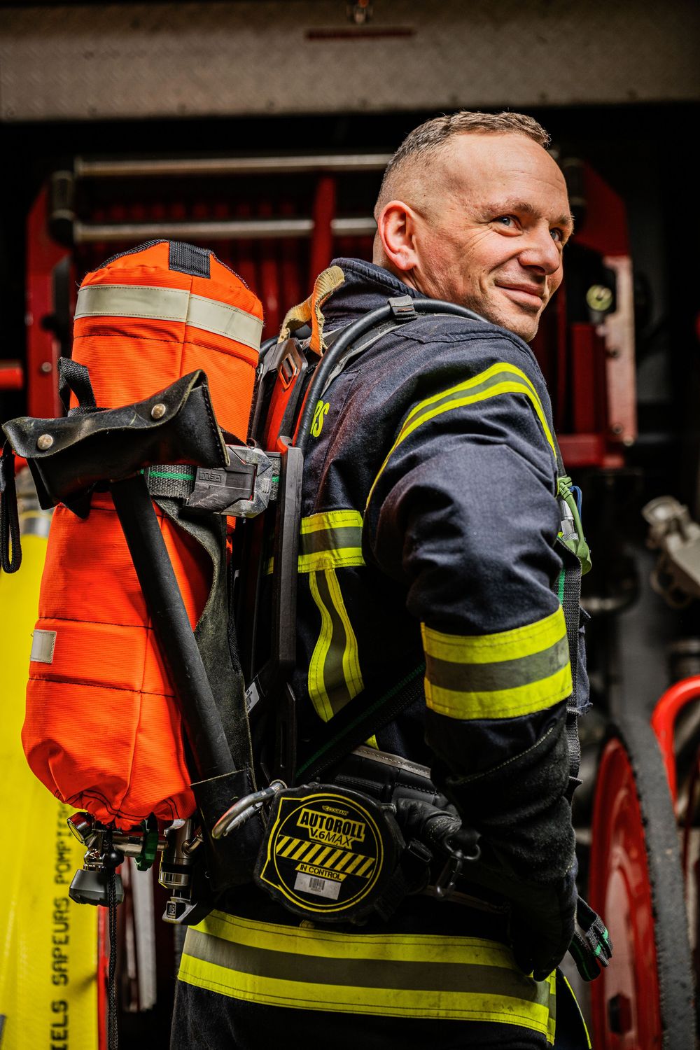 A male firefighter in reflective gear carries an oxygen tank while standing beside a fire truck.