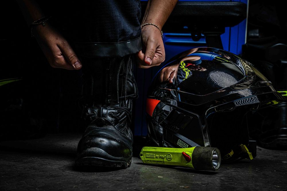 Firefighter prepares gear beside motorcycle boots and yellow helmet on dark floor.