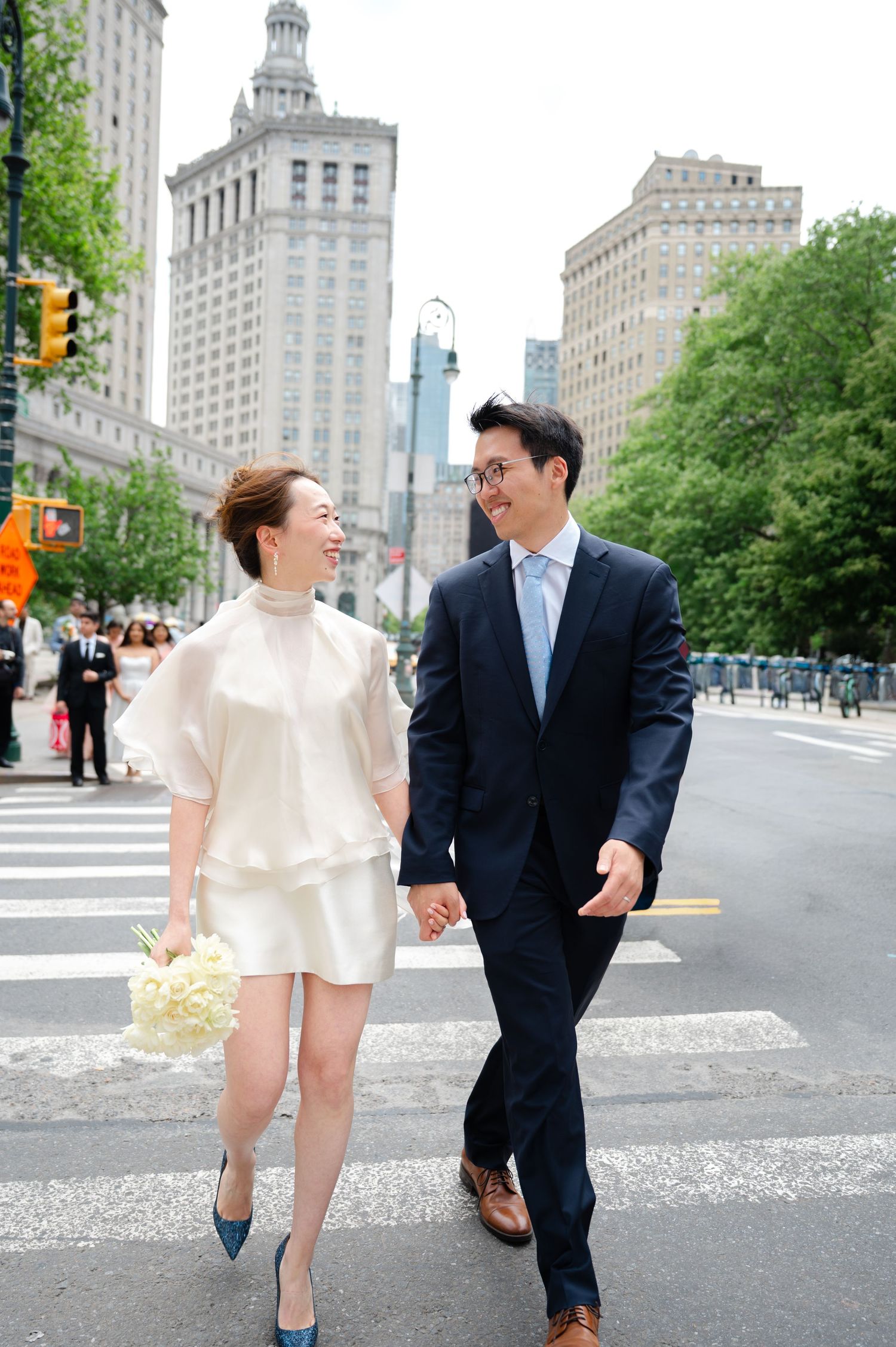 Couple in wedding attire holding hands crossing a city street in New York, woman in white mini dress holding bouquet before eloping.