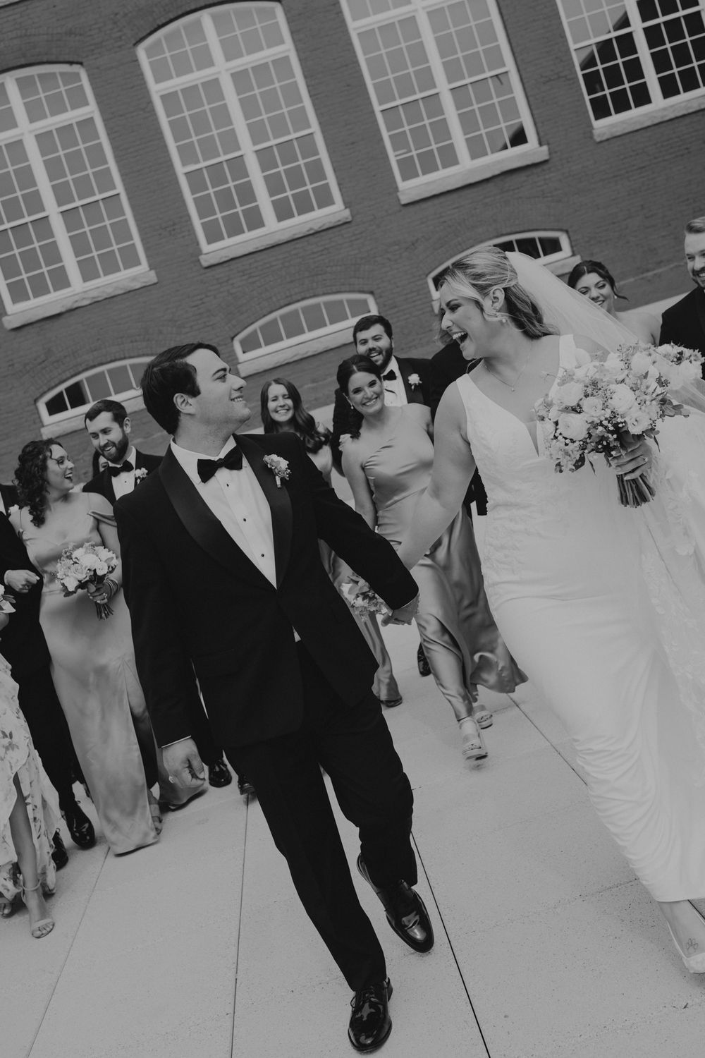 Black and white photo of a smiling bride and groom walking hand in hand with their wedding party outside a brick building.