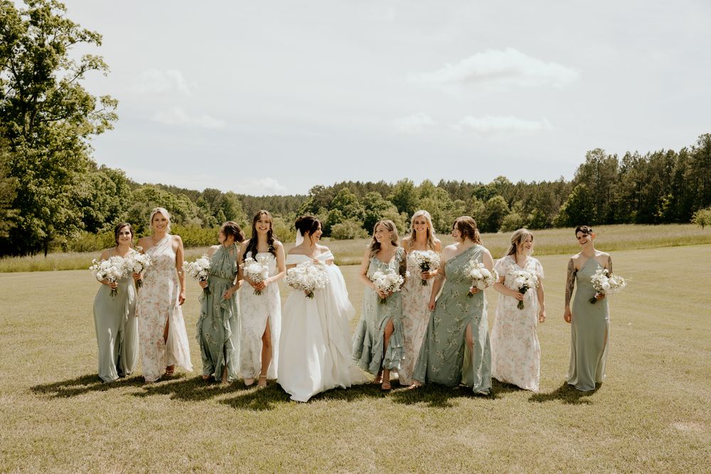 Bride and bridesmaids in sage green gowns walk across a lush green field on a sunny wedding day.