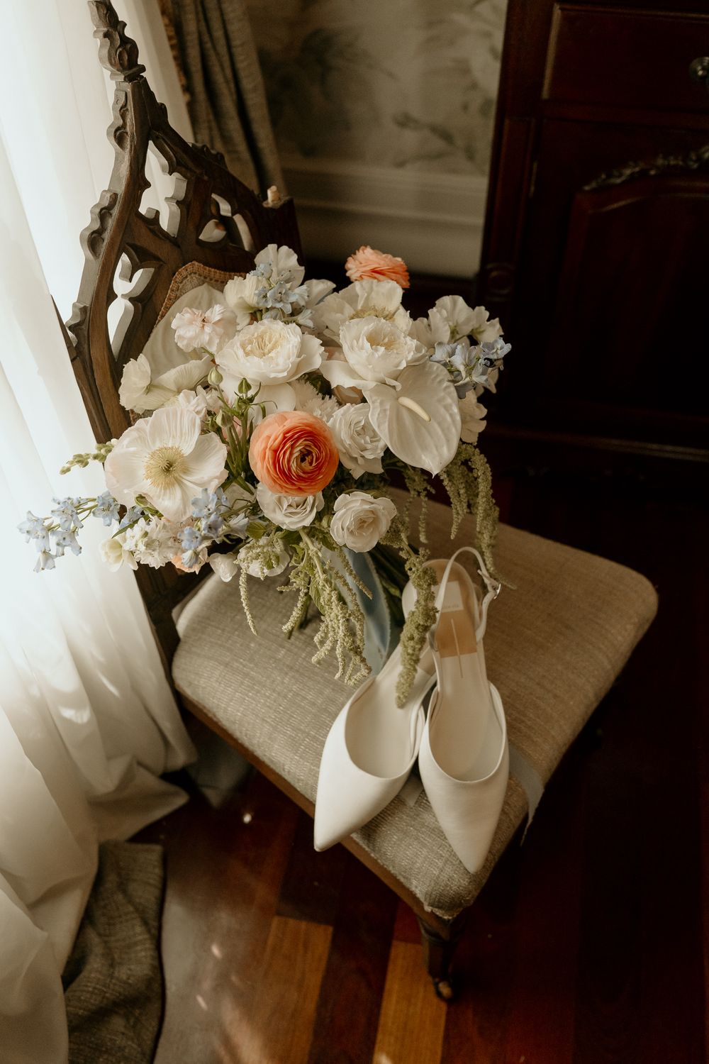 White bridal heels and a bouquet of white, peach, and blue flowers rest on a vintage wooden chair near a window.