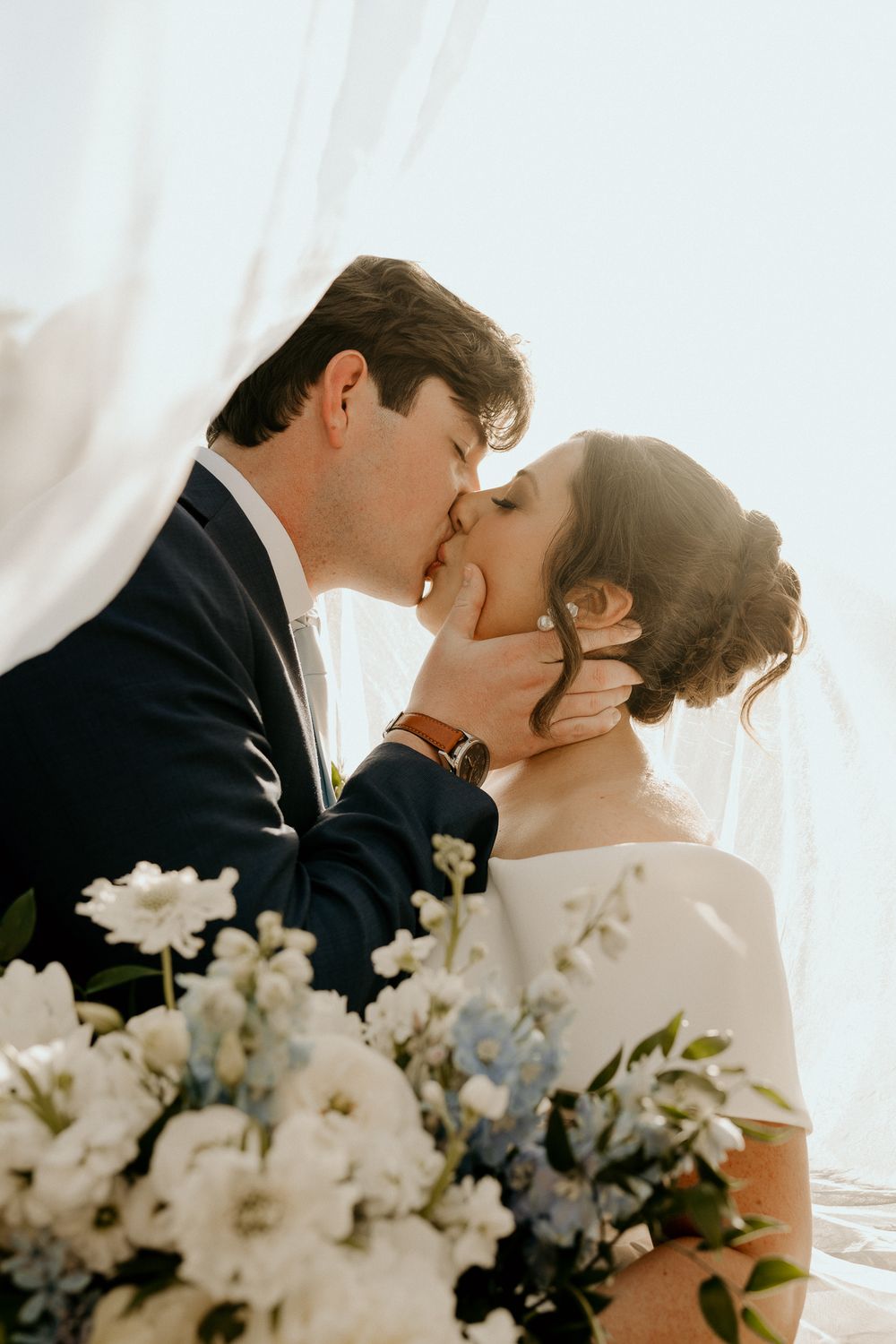 A bride and groom share a romantic kiss, with a white floral bouquet in the foreground and soft backlit light.