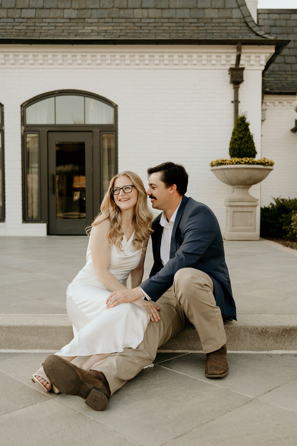 A smiling couple sits on stone steps outside a white brick building, the woman in a white dress and the man in a navy blazer.