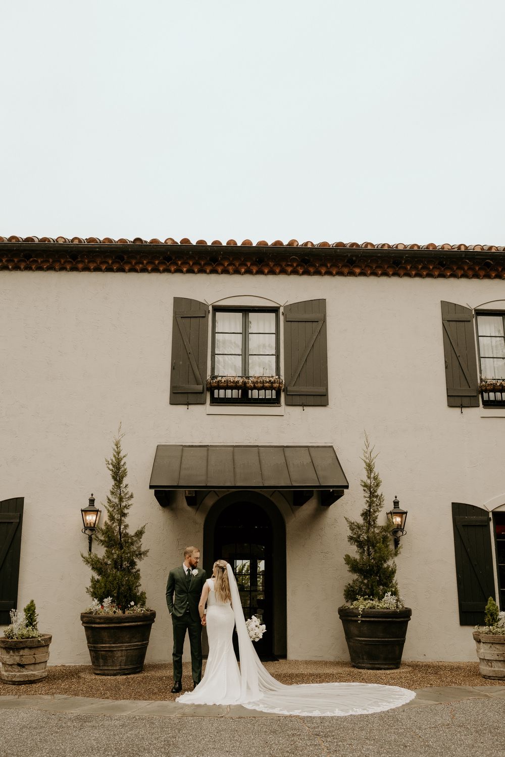 Bride and groom stand before elegant Spanish-style venue with dark shutters, arched entry, and tall potted topiaries on their wedding day.