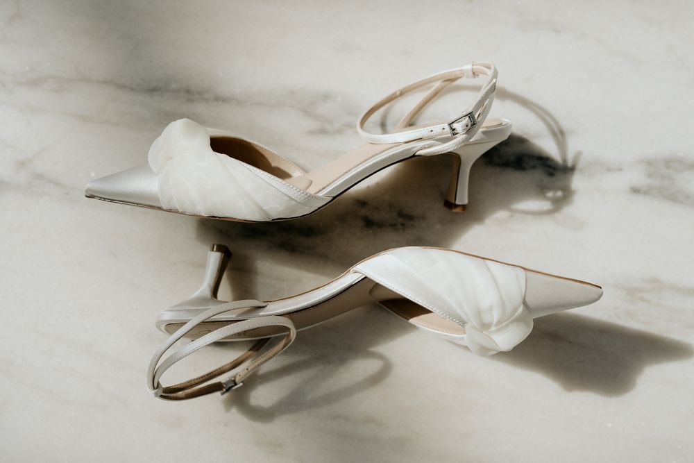 Elegant white satin kitten heel sandals with ankle straps displayed on a marble surface.