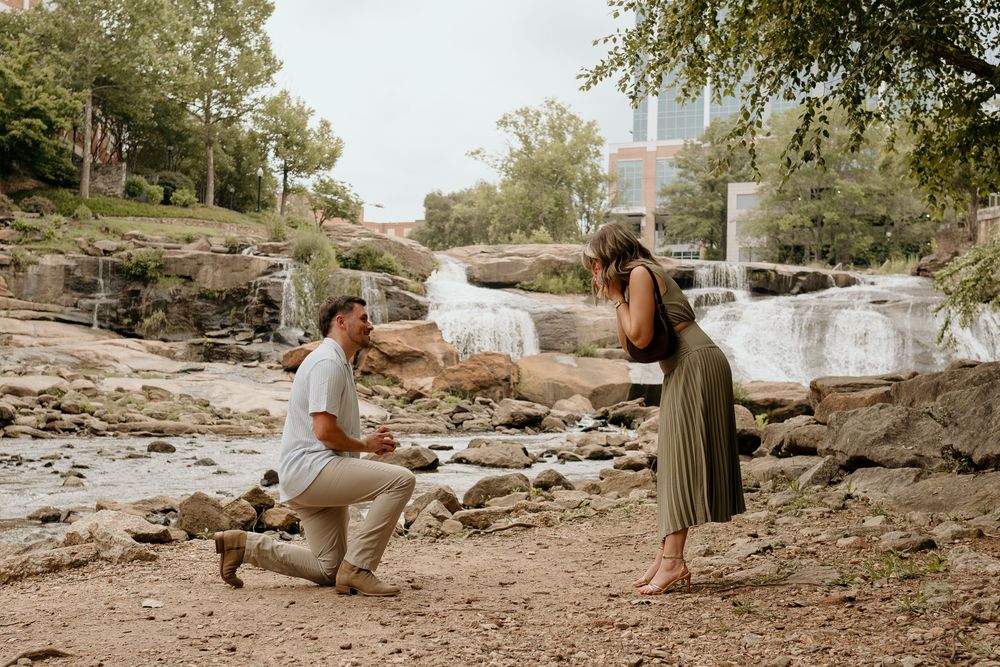 Man proposing to woman on one knee near a waterfall in an outdoor park setting on a sunny day.