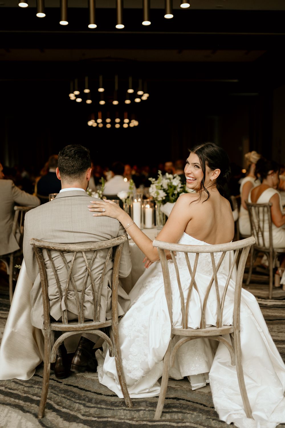 Bride in white strapless gown smiles back at camera while seated with groom at elegant wedding reception.