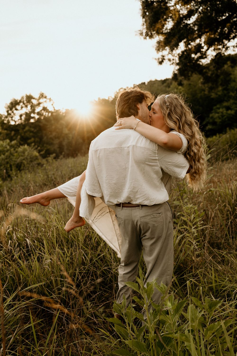 A couple shares a romantic kiss at golden hour in a lush green field, as the man lifts the woman in his arms.