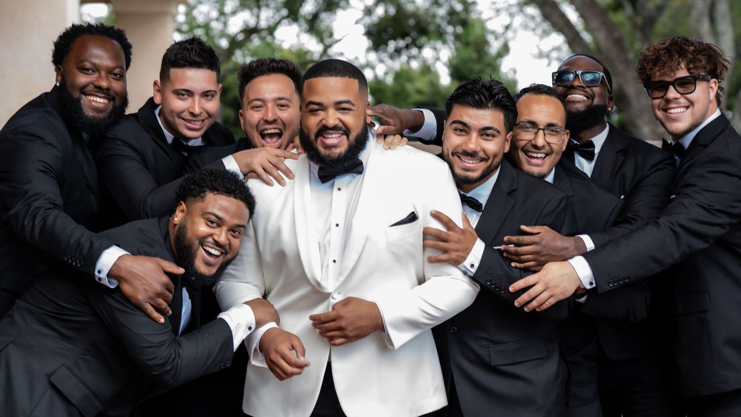 Groom in white tuxedo jacket celebrates with groomsmen in black suits at outdoor wedding ceremony.