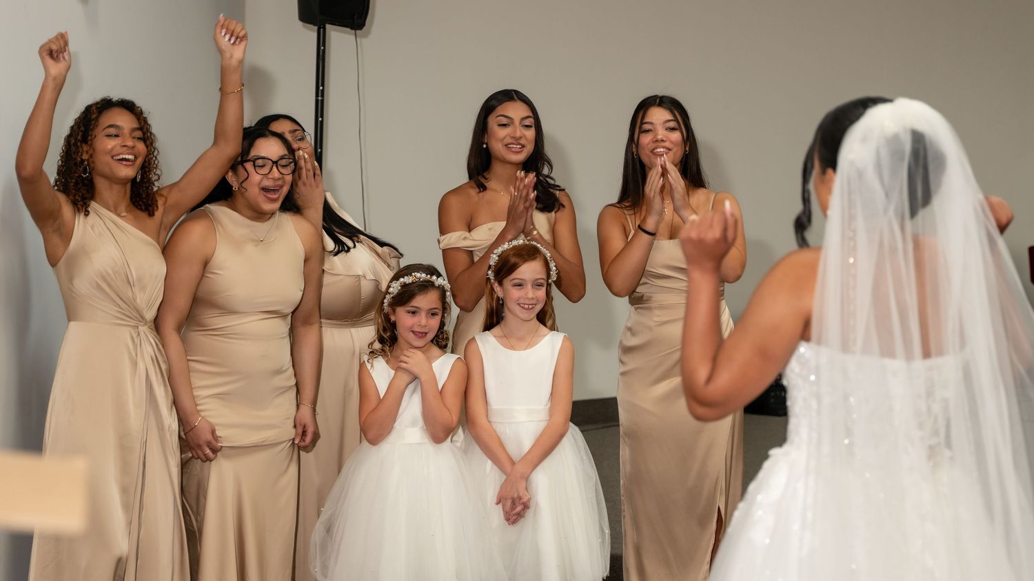 Bridesmaids in champagne gowns and flower girls react with joy as bride in white veil reveals her wedding look.