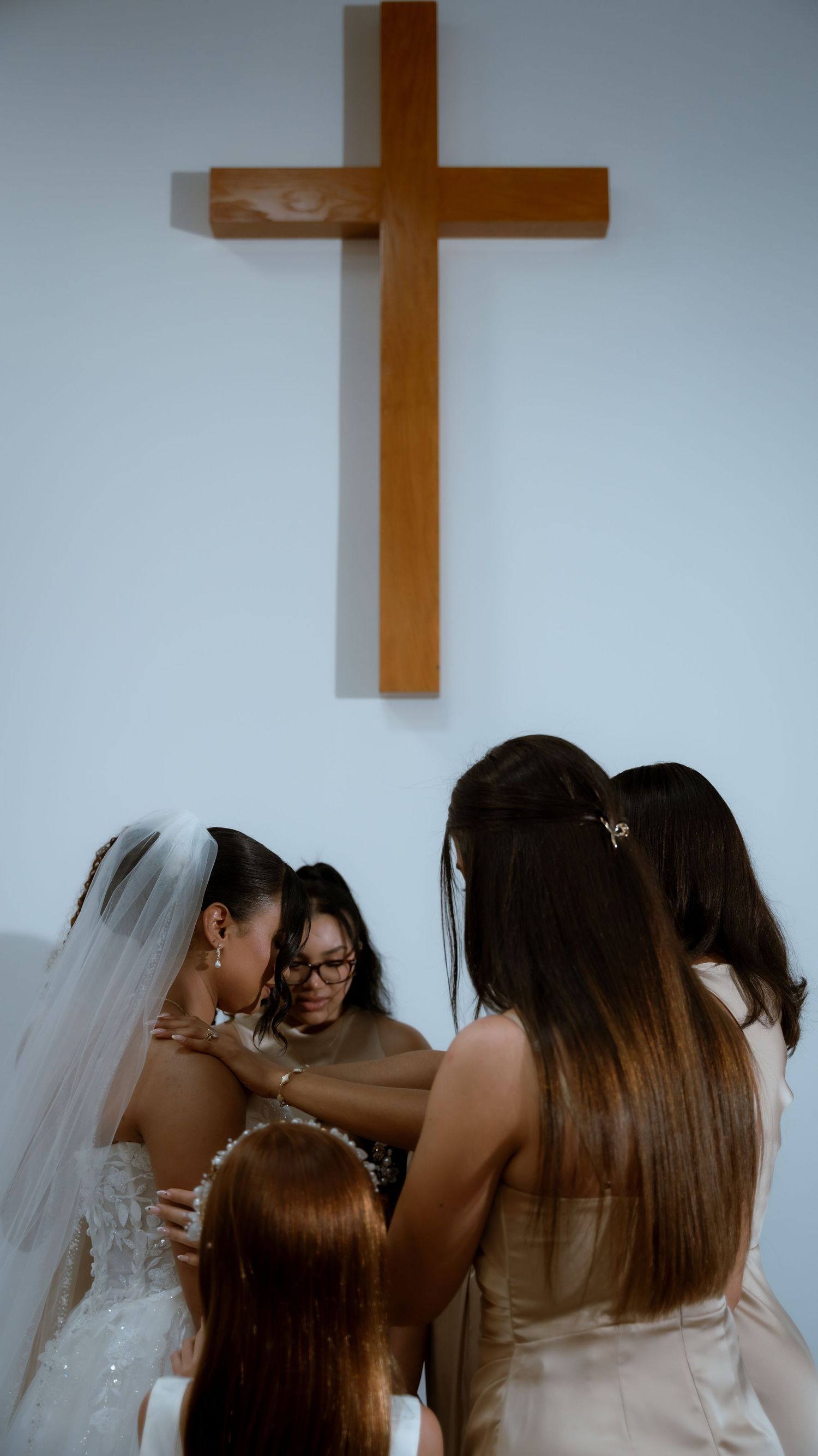 Bride surrounded by bridesmaids in emotional moment under wooden cross in church.