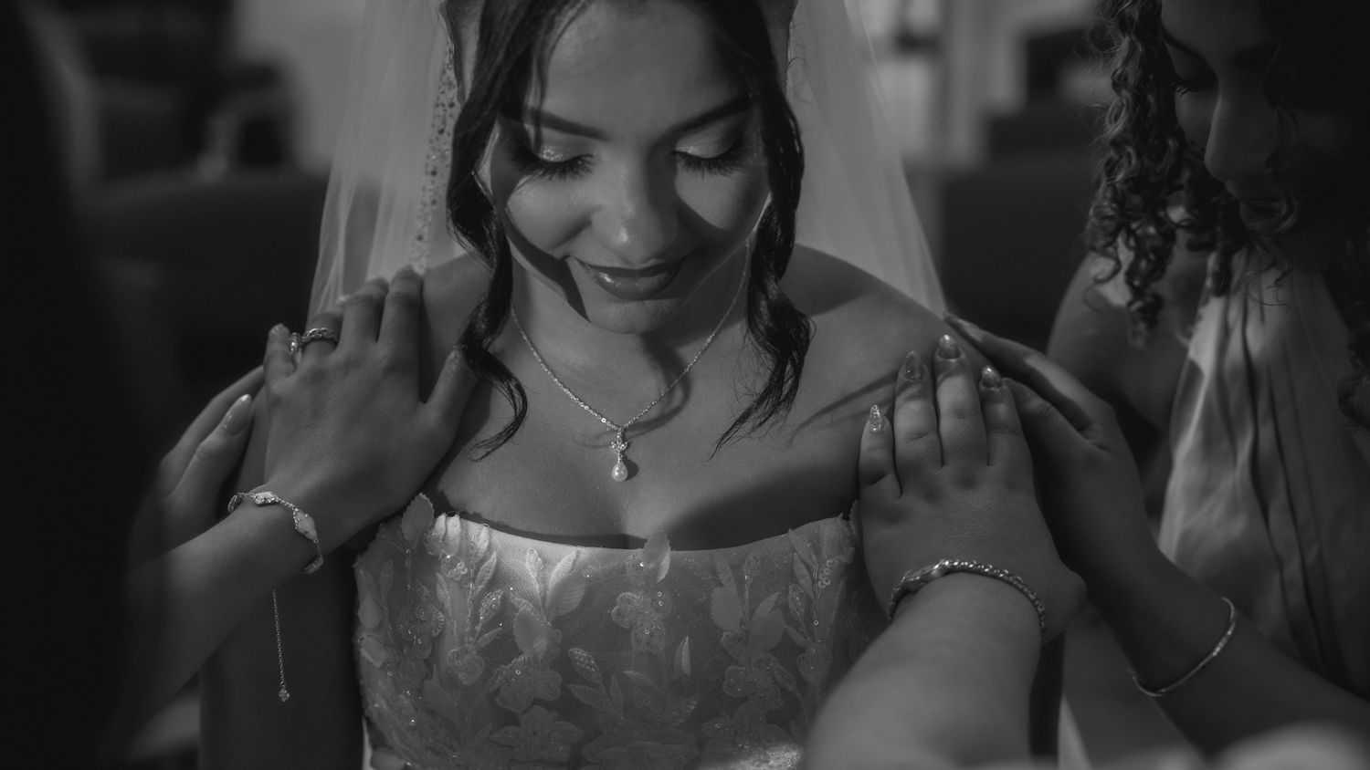 Black and white photo of a smiling bride in a lace gown and veil, being helped by attendants on her wedding day.