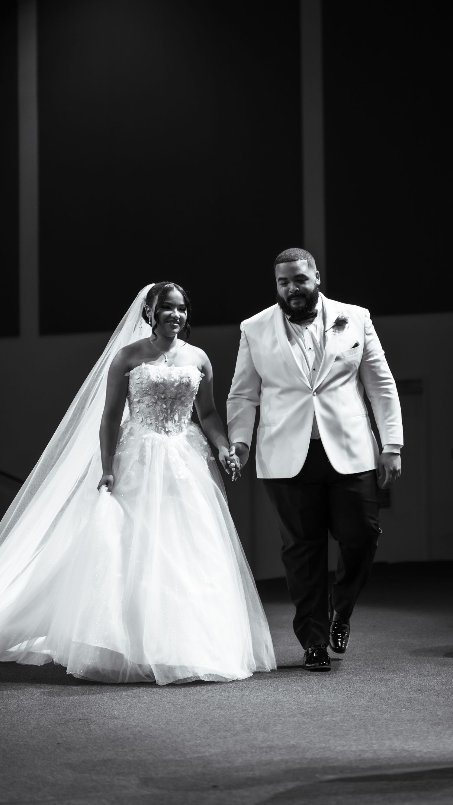 Newlyweds walk hand in hand in black and white wedding portrait.
