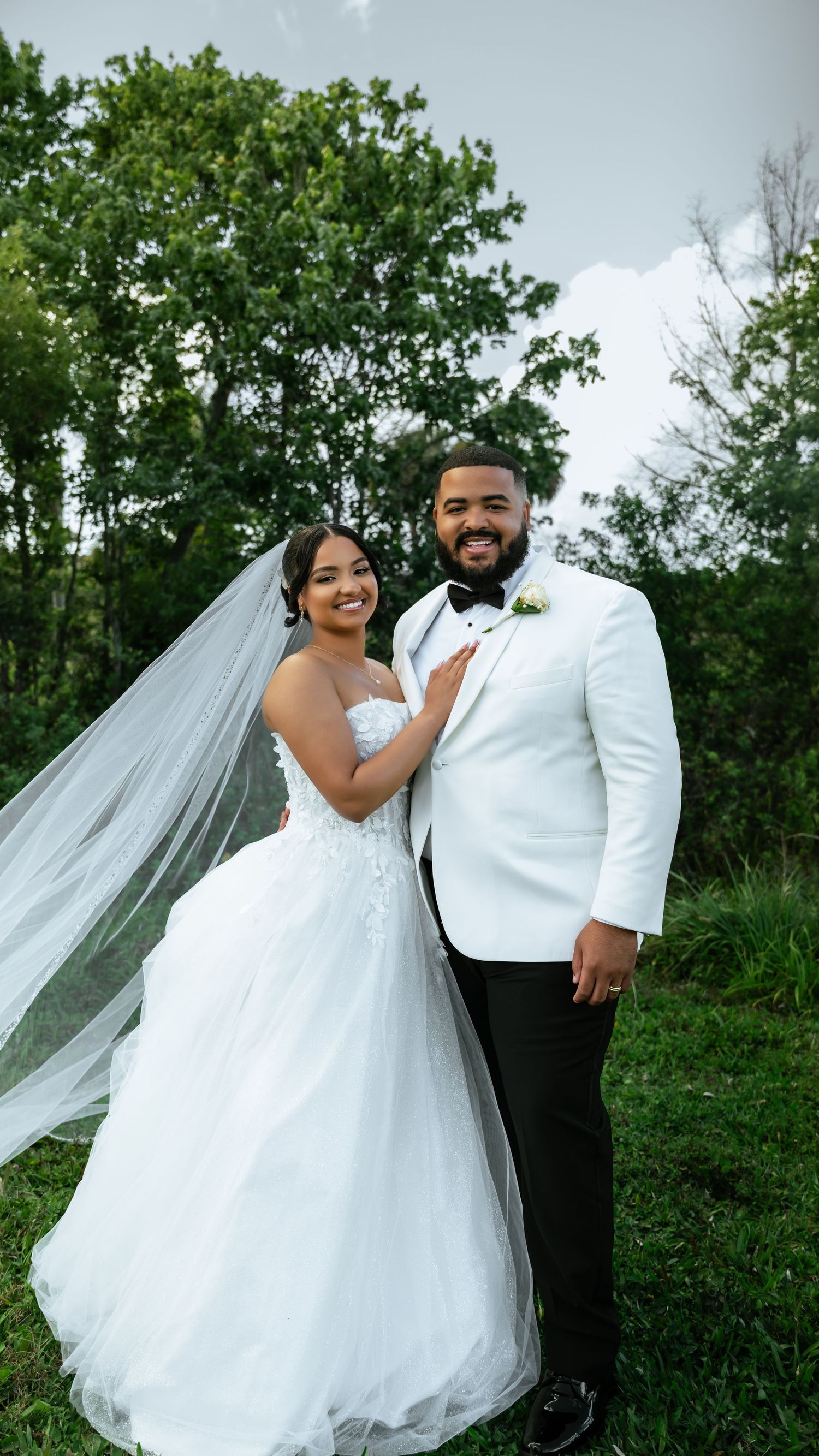 Bride and groom posing outdoors in wedding attire, she in a white gown with veil, he in a white tuxedo jacket.