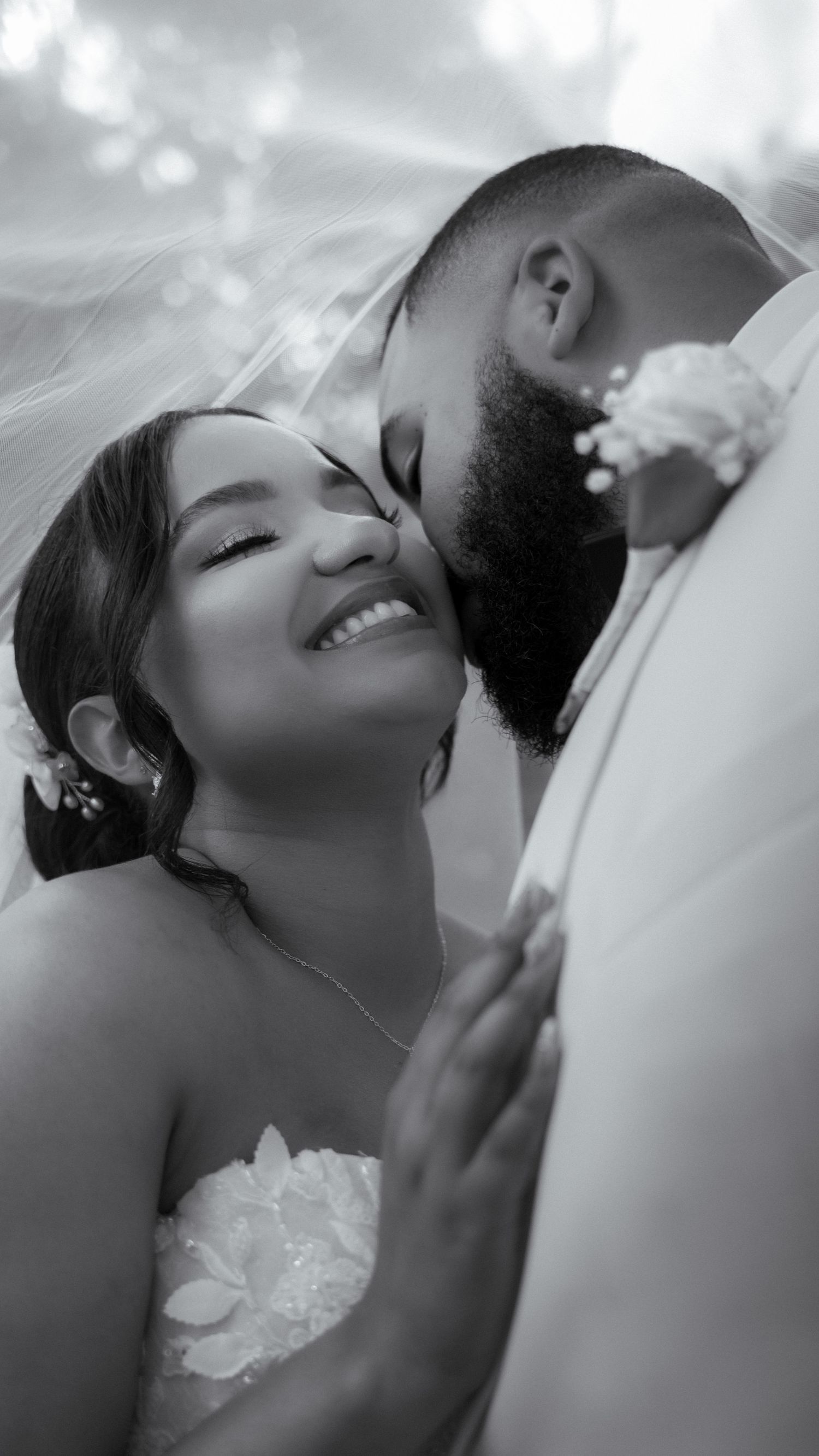 Black and white wedding photo of smiling bride under veil, groom kissing her cheek, romantic couple portrait.