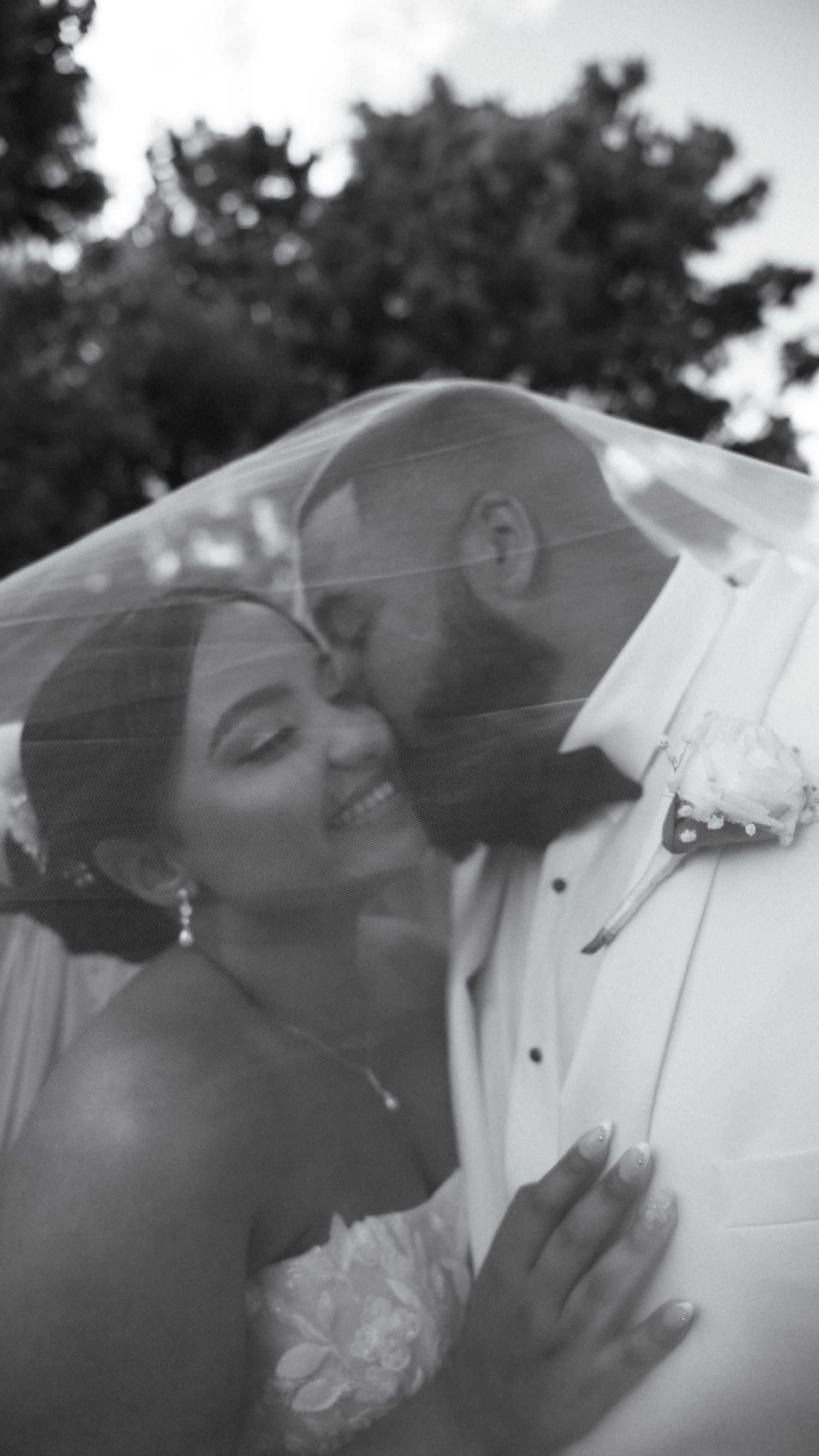 Black and white wedding photo of groom kissing bride on cheek under veil outdoors.