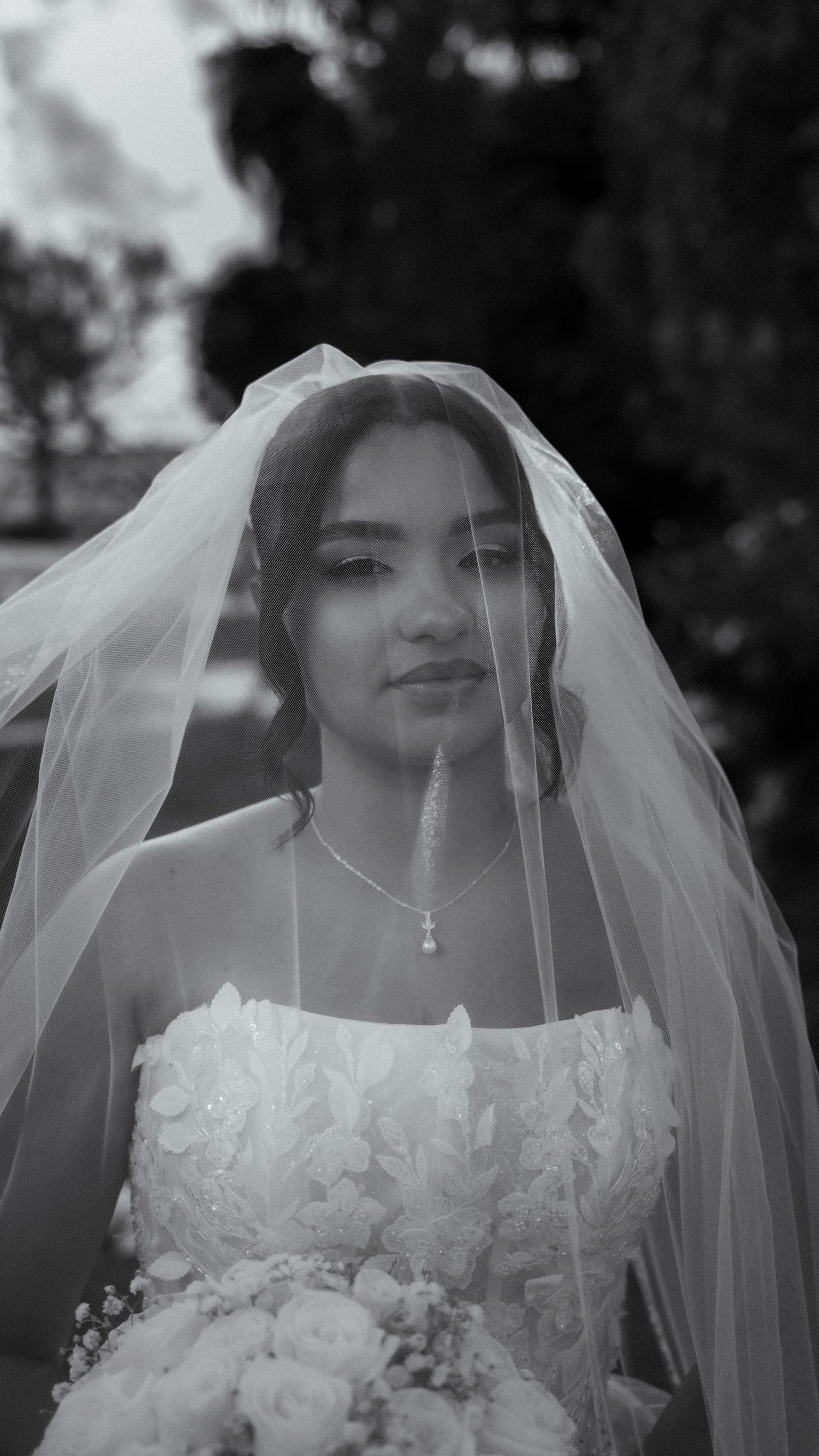 Elegant bride in white gown and veil holding bouquet in black and white portrait.