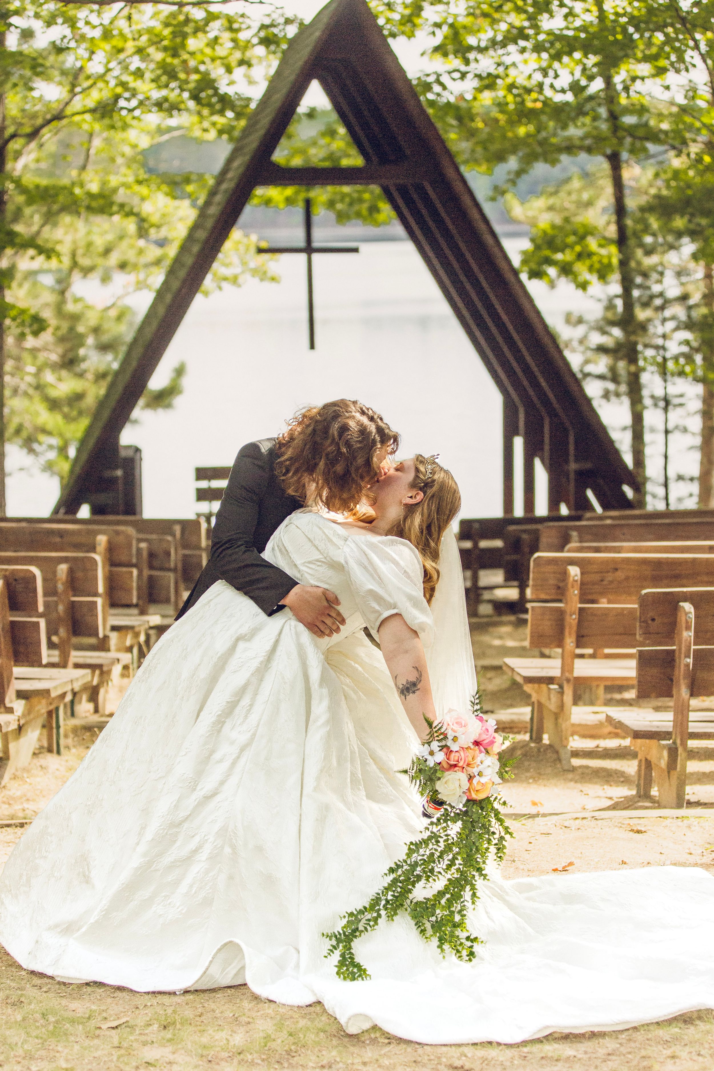 Newlywed couple sharing a romantic kiss in an outdoor chapel with wooden pews, cross, and lush trees in background.